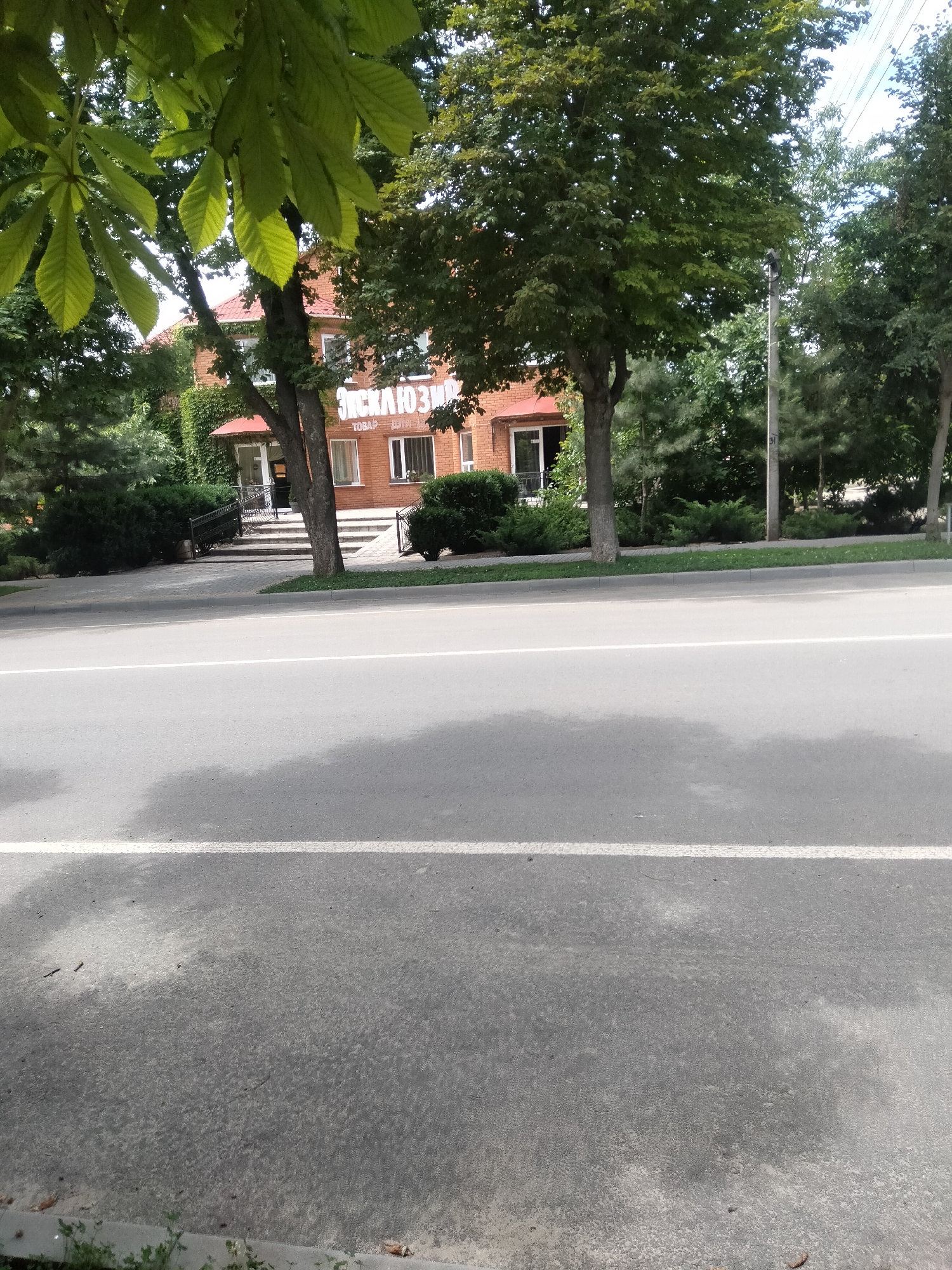 The image shows a building with an entrance and a sign that reads "ЭКСКЛЮЗИВ". The building is made of brick and has a red roof. It is surrounded by trees and bushes. A road with a white line runs in front of the building. A person can be seen walking up the stairs to the building's entrance. The scene appears to be a commercial establishment in a semi-urban or suburban area.