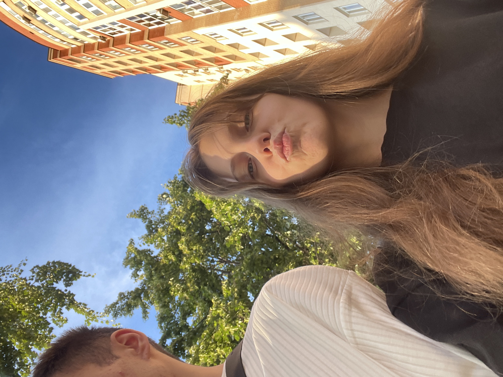 In the city of Ufa, Russia, an outdoor selfie is taken. A young woman with light brown hair is in the foreground, puckering her lips. She is wearing a black t-shirt. To her left, a young man's head and shoulder are visible; he is wearing a white, ribbed shirt. Behind them, lush green trees are illuminated by the sun, and a tall, modern apartment building with orange and beige facades stands against a clear blue sky. The image captures a moment of youthful outdoor activity.