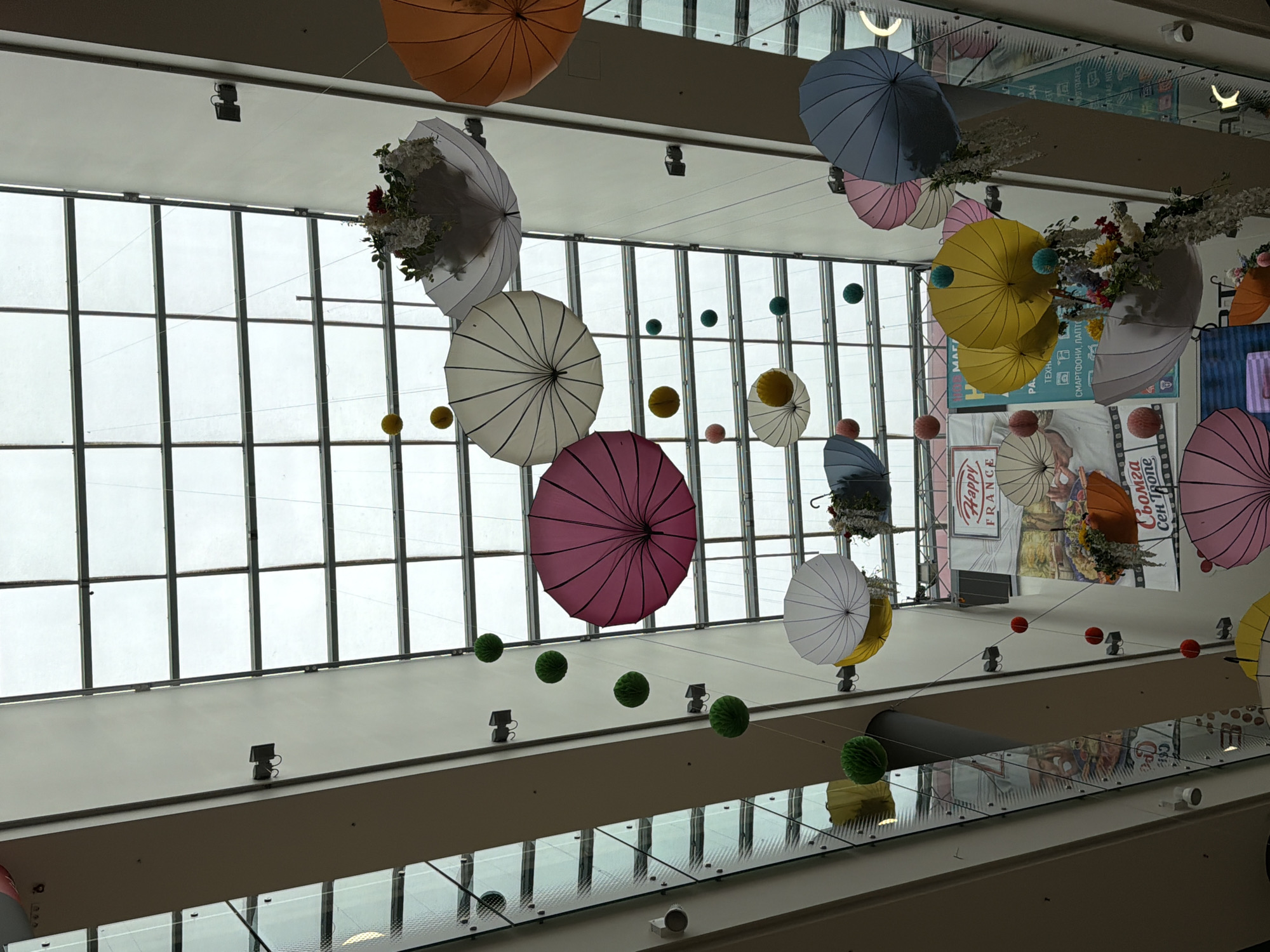 The image shows an upward view of a shopping mall ceiling in Unknown, Unknown, decorated with colorful umbrellas and spheres suspended on wires. The ceiling has a grid-like structure of windows, allowing natural light to filter through. Various spheres in green, yellow, and red are hung at different heights. The umbrellas are a mix of colors, including orange, white, pink, yellow, and blue, some filled with floral arrangements. The mall's architectural elements include balconies with glass railings and recessed lighting. A vertical advertisement is visible through the windows, further suggesting the commercial nature of the space.