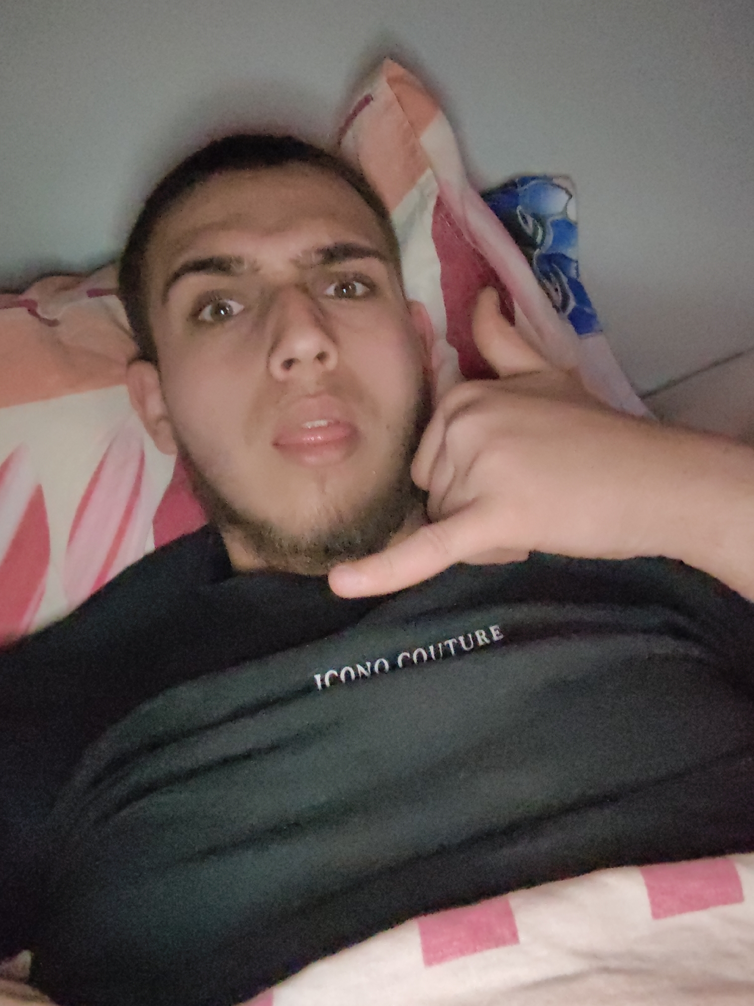 A young man is lying down, possibly in bed. He is wearing a dark shirt with "ICONO COUTURE" printed on it. He is making a "call me" gesture with his hand. He has short dark hair and a beard. The background includes bedding with colorful patterns. There is no information in the image to suggest a specific location in Sofia, Bulgaria.