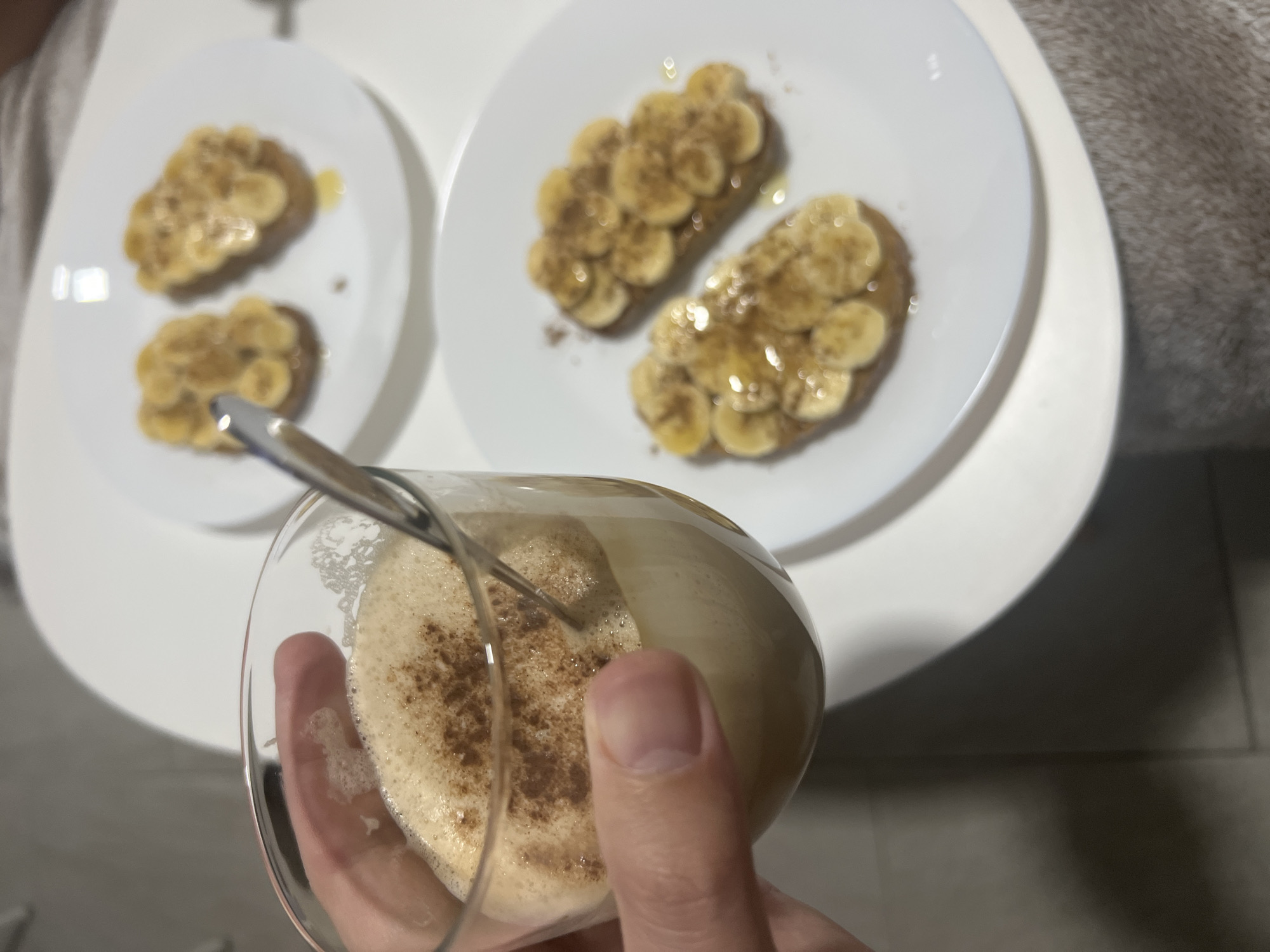 A first-person shot shows a hand holding a glass of foamy, light brown liquid, possibly a cappuccino, with a silver spoon inserted. The liquid is topped with a sprinkle of dark brown powder. The background features two white plates, each containing two pieces of toast topped with sliced bananas and a drizzle of sauce. The setting is a white table, with a glimpse of a gray garment and a tiled floor in the periphery. The location is potentially Mogán, Spain, based on the user's request, but there are no specific clues visible in the image to confirm this.