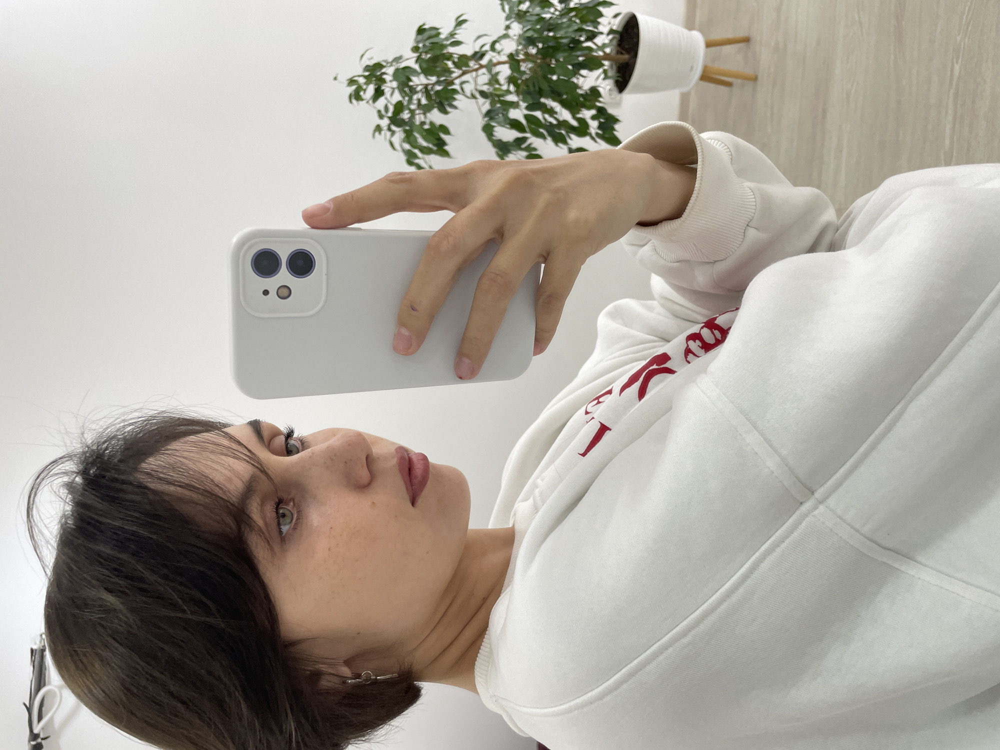 The image, taken in Izhevsk, Russia, captures a young woman taking a selfie or recording a video with a light-colored smartphone. She is viewed from a side profile, her gaze directed towards the phone screen, which is out of frame. She has dark hair, styled with bangs, and light-colored eyes. Her complexion shows a scattering of freckles, and she wears light lipstick.
She is dressed in a white hooded sweatshirt, or hoodie, which features a prominent, but partially obscured, red design on the front chest area. Her right hand holds the light-grey or white smartphone, encased in a protective cover, with its dual camera system and flash visible on the back. Her left hand is not visible.
The background is minimalist, consisting of a plain white wall on the left side of the frame. To the right, a potted green plant in a white ceramic pot sits on a light-colored wooden stand. The floor behind the plant appears to be light-toned, possibly wood or laminate. The lighting is soft and even, typical of an indoor environment. The woman's expression is neutral and focused on the activity of using her phone. The red design on her hoodie is not legible as specific text.