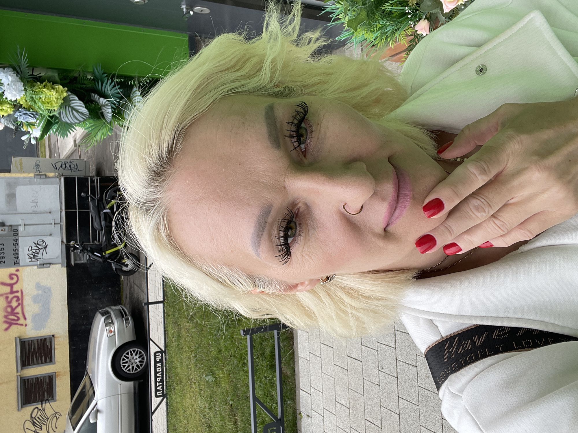 The image shows a close-up selfie of a blonde woman with green eyes and prominent false eyelashes. She is wearing a white jacket and has bright red fingernails. A nose ring is visible, and she is touching her chin with her right hand. Behind her, a silver car is parked next to a building with graffiti. A black scooter is also visible. To the right, there is a green wall and some artificial greenery with flowers. The scene appears to be an outdoor urban setting. The graffiti on the building includes the word "YORSH" in large pink letters. The license plate on the car is not clearly visible. The overall impression is a snapshot taken in an urban environment in Saint Petersburg, Russia, given the graffiti styles and general urban landscape which are consistent with the city.