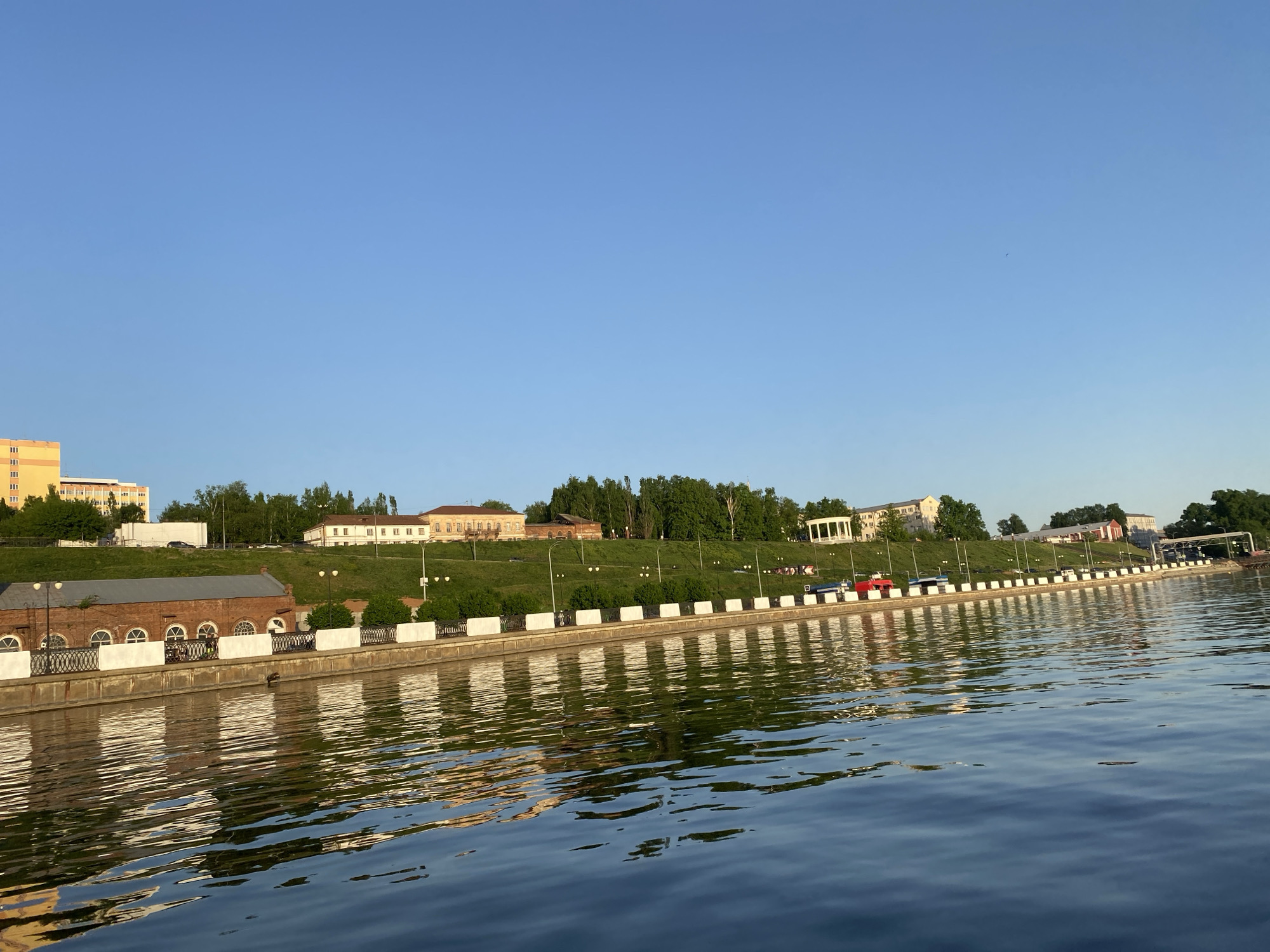 The image shows a waterfront scene in Izhevsk, Russia. The foreground is dominated by the dark blue, rippling water of the Izhevsk Reservoir. A concrete and brick embankment runs along the shore. A low brick building with arched windows is visible on the left, followed by a series of white bollards and decorative iron fencing. Beyond the embankment, a grassy slope rises toward various buildings, including a yellow multi-story building and other structures. Trees are interspersed among the buildings. The sky is clear and blue. Several cars are parked at the end of the esplanade. The perspective suggests the photo was taken from a boat or floating platform on the water. The time of day is likely late afternoon or early evening, judging by the light.