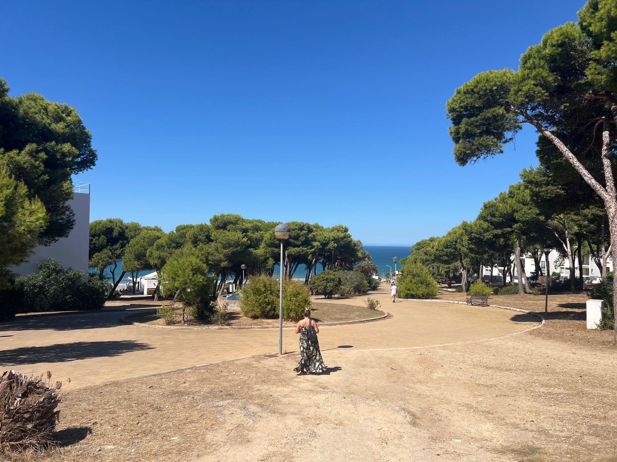 The image depicts a sun-drenched, outdoor scene, likely in Conil De La Frontera, Spain, judging by the coastal vegetation and architecture. The foreground is dominated by dry, sandy ground with some sparse, dead vegetation. A paved walkway, a light tan color, curves through the middle ground. A woman in a long, patterned dress walks away from the camera on this path, casting a shadow. She has dark hair and carries a black backpack. To the left of the path, there is a white building with a railing on its roof, partially obscured by tall, green pine trees. Beyond the walkway, more pine trees and some bushes create a park-like setting. In the background, the blue ocean is visible, with a hint of a beach and a few white structures that could be umbrellas or small buildings. Two lampposts are present in the mid-ground. The sky is a clear, bright blue. To the right, further back on the path, a person in white clothing is walking towards the left. The overall impression is one of a warm, sunny day in a coastal resort area.