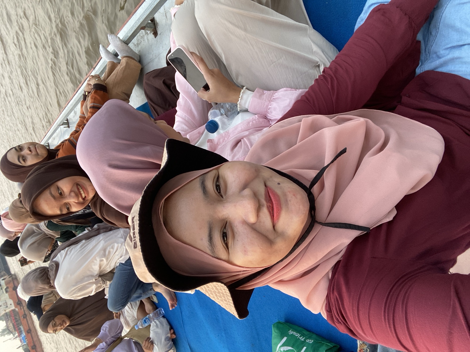 The image shows a group of people, predominantly women wearing hijabs, sitting on a boat in what appears to be a river. The boat has a blue mat covering its floor. The people are casually dressed and some are looking at the camera, while others are engaged in their own activities. One woman in the foreground is taking a selfie, smiling and wearing a straw hat. Another woman is holding a smartphone. In the background, other boats and the water of the river are visible. The lighting suggests it might be late afternoon or early evening. The setting is consistent with a riverine environment possibly in a region like Sungai Pitung, Indonesia, where boat travel is common.