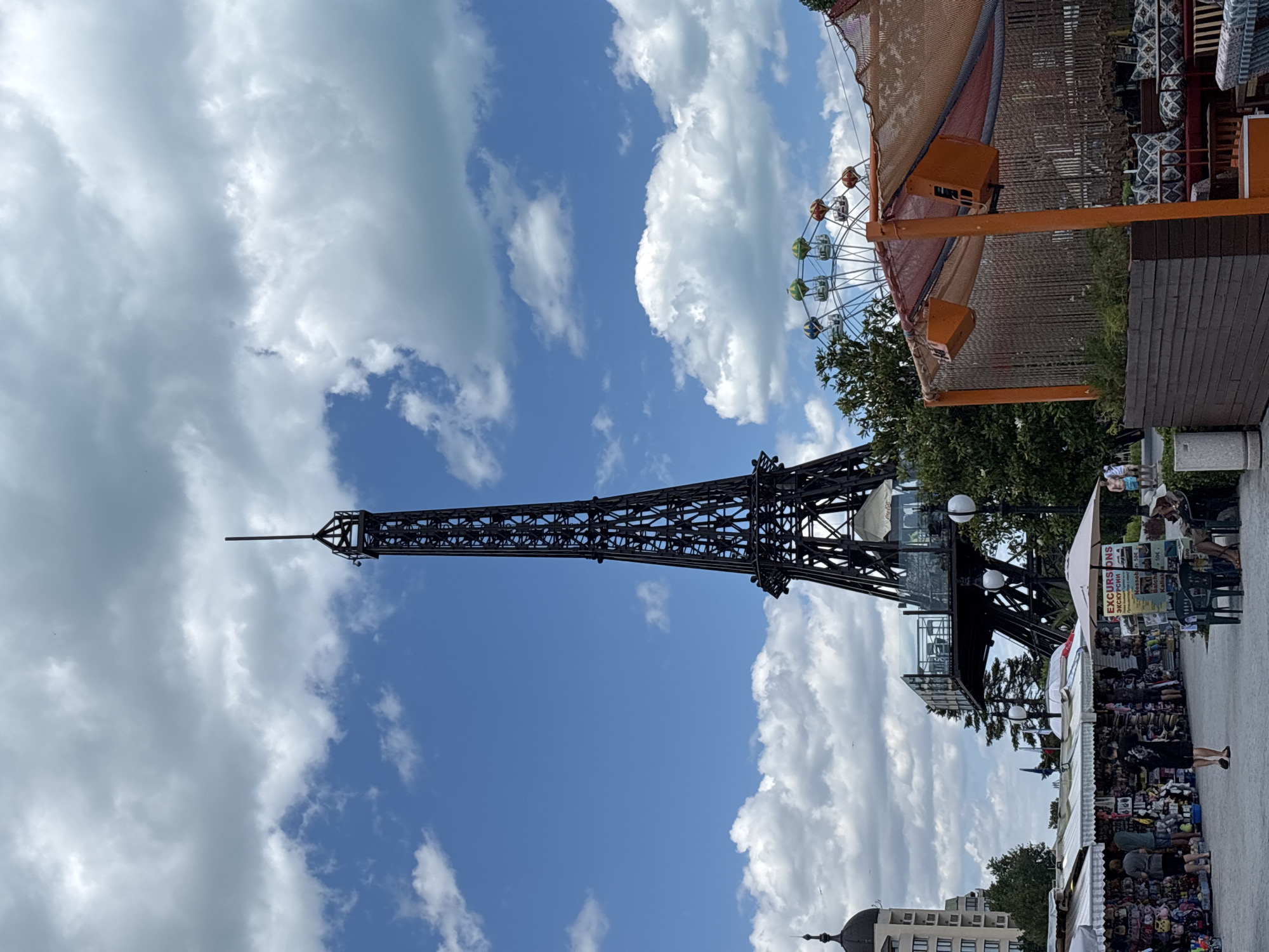 The image shows a miniature replica of the Eiffel Tower in Varna, Bulgaria. The tower is made of black metal and has a viewing platform partway up. In the background, a Ferris wheel can be seen, suggesting an amusement park or entertainment area. In the foreground, there are market stalls selling various goods, with people browsing and shopping. The scene appears to be a lively public space, possibly a tourist attraction. The sky is blue with scattered clouds, indicating fair weather.