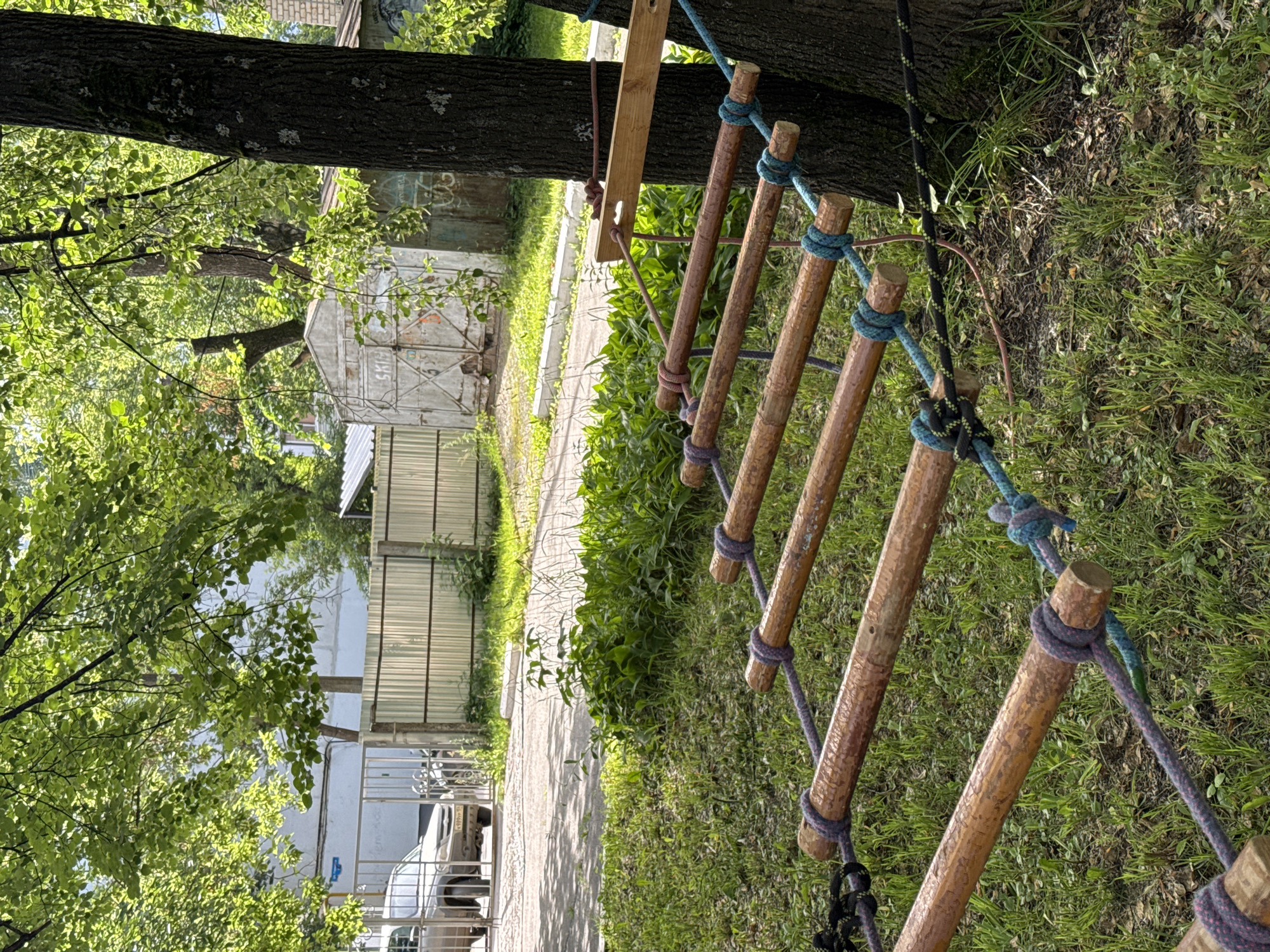 A rope ladder made of wooden rungs tied with colorful ropes is laid out on a grassy area in Chaikovsky, Russia. The ladder is attached to a large tree on the right side of the image, and another rope is tied to a wooden plank also attached to the tree. In the background, a metal fence, a car, and a garage are visible. The scene is bathed in natural sunlight.
