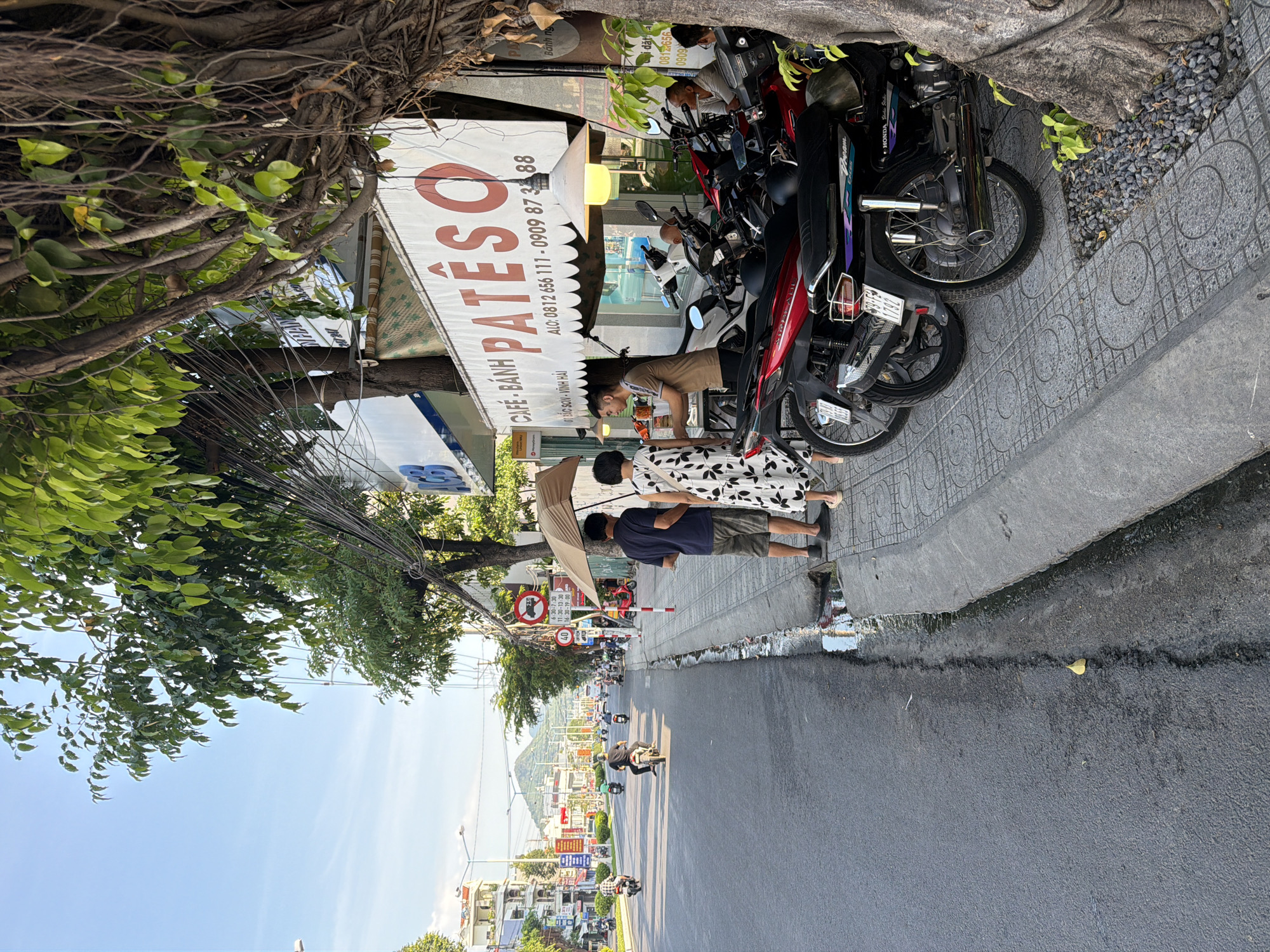 The image depicts a street scene in Bac Nha Trang Ward, Vietnam. A paved road runs along the left side of the frame, with a sidewalk on the right. Several motorcycles are parked on the sidewalk. A cafe or shop with a sign that reads "CAFE - BANH PATESO" is visible, with a large lampshade hanging in front. Three people are standing on the sidewalk near the cafe: a man in a dark blue shirt and shorts, a woman in a white dress with a floral pattern, and another man in a light brown shirt. The man in the brown shirt appears to be interacting with the woman in the floral dress. The woman is holding a beige umbrella. Further down the street, more motorcycles and people can be seen, along with buildings and trees lining the road. The sky is clear and blue.