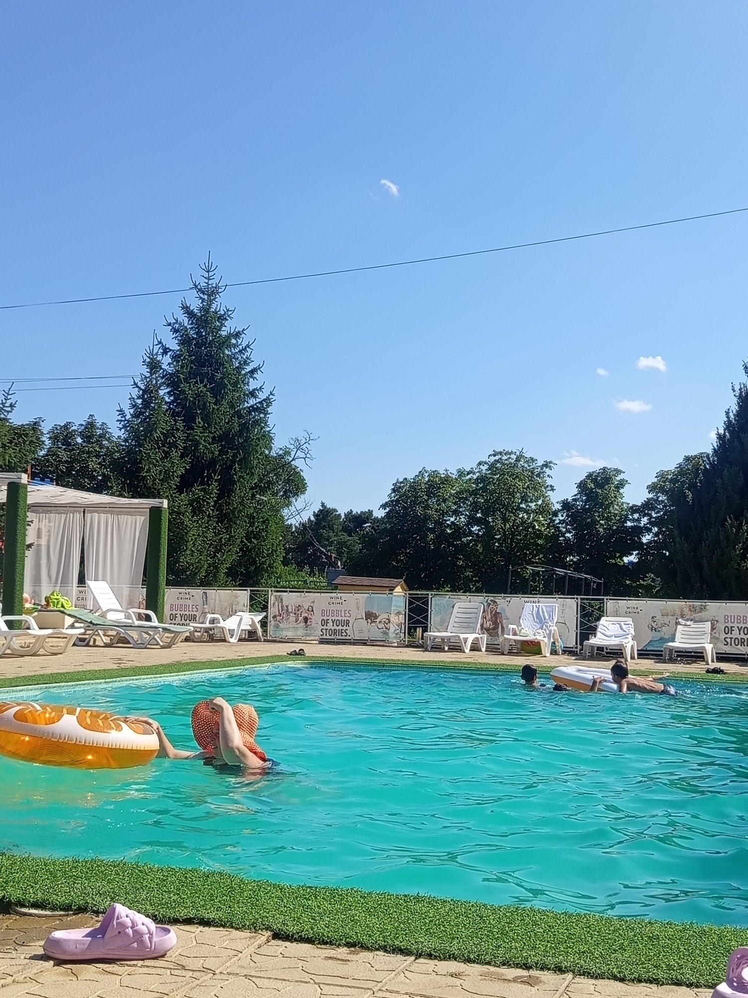 In Molovata Nouă, Moldova, a bright, sunny day is captured at an outdoor swimming pool. The turquoise water is clear and inviting, with several people enjoying the refreshing dip. In the foreground, a person wearing a wide-brimmed orange hat is partially submerged, holding onto an inflatable ring. Further in the pool, two children are swimming. The pool area is bordered by a vibrant green artificial turf, and beyond that, a tiled patio with several lounge chairs arranged on it. In the background, mature trees and a white gazebo add to the serene atmosphere. A banner with "WINE CRIME BUBBLES OF YOUR STORIES" is visible behind the lounge chairs, suggesting a place that combines leisure with perhaps a vineyard or winery. The sky is a clear, brilliant blue, indicating a perfect summer day.
