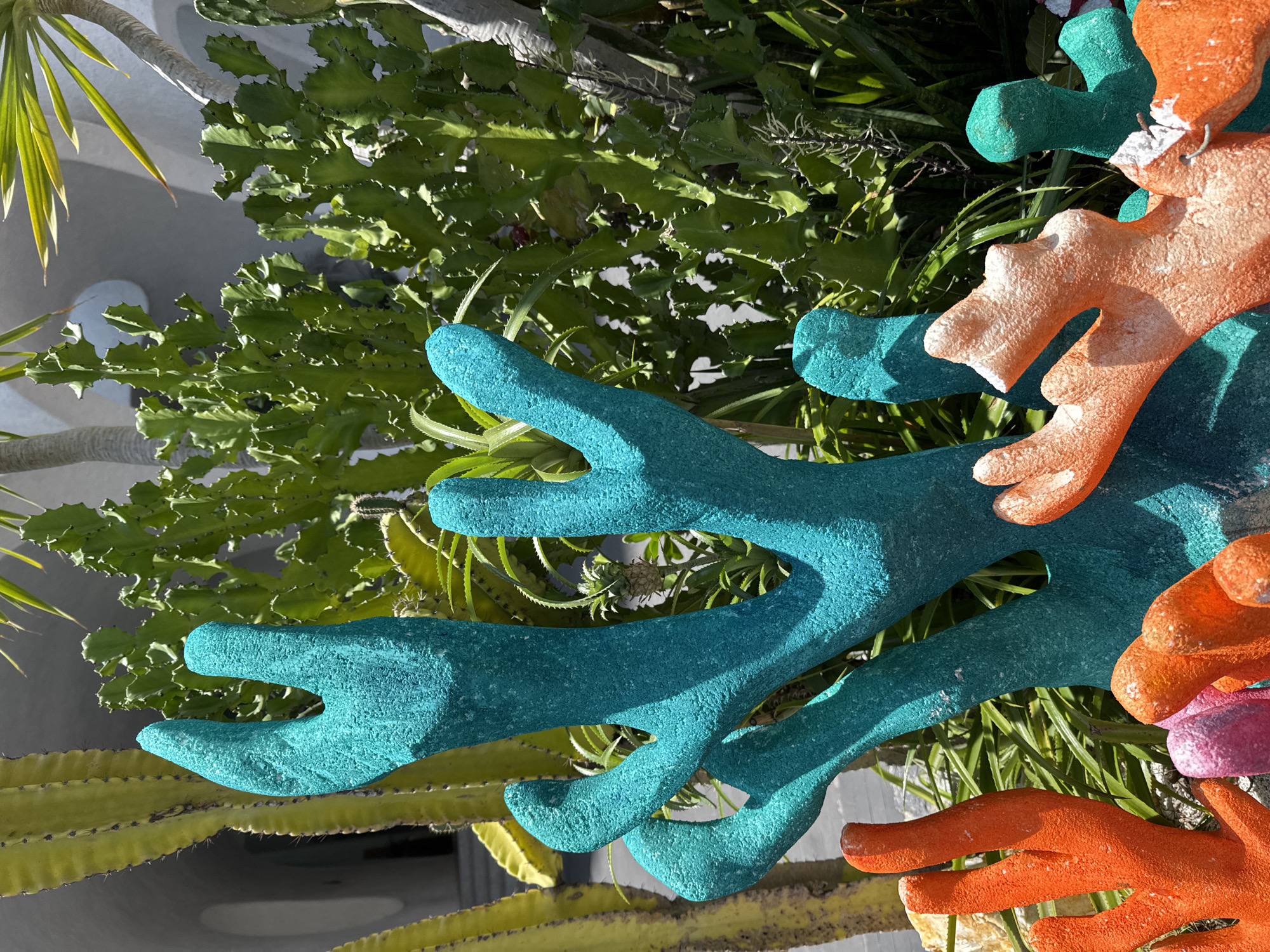 The image displays a close-up view of brightly colored, artificial coral sculptures in an outdoor setting. The sculptures are vibrant shades of teal, orange, and pink, with a rough, textured surface. In the background, various types of cacti and other succulents are visible, along with architectural elements that suggest a modern building with curved, white surfaces. The lighting indicates a sunny day, casting shadows on the sculptures and plants. This scene is located at the VinWonders Nha Trang amusement park on Hon Tre Island, Nha Trang, Vietnam, specifically within the Mermaid Lagoon area, designed to evoke an underwater theme. There are no people or animals visible in the frame.