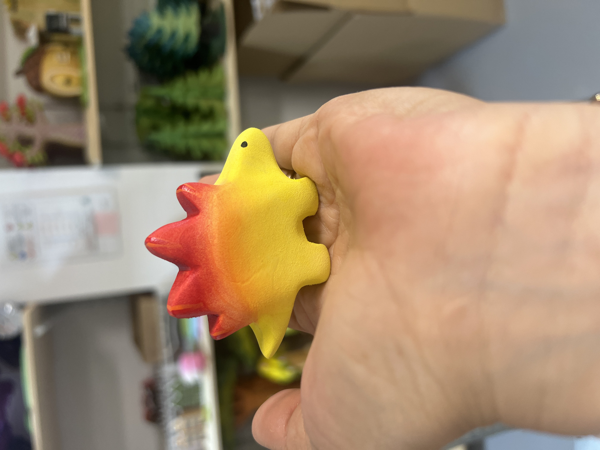 A person's hand is holding a star-shaped object that is yellow with a gradient of red on some of its points. The object has a small black dot on one of its points, resembling an eye. The background is blurred and shows what appears to be a store shelf with various items, including some green, spiky objects and a light brown object with a face. The lighting suggests it is daytime.