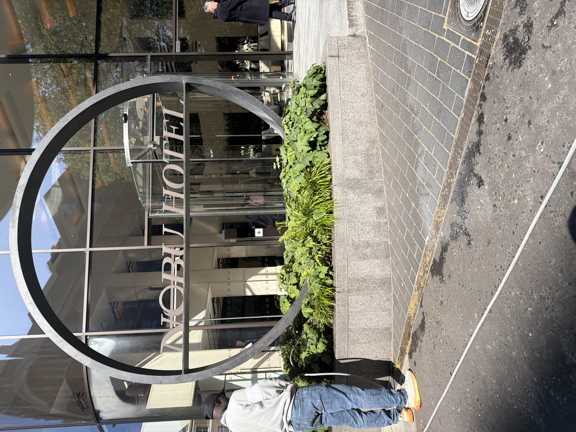 The image shows the exterior of the Nobu Hotel in London, United Kingdom. A large, circular metal sign displays the hotel's name. The hotel entrance features glass doors and windows. Landscaping includes green plants in a raised planter adjacent to the entrance.
A man stands near the planter, wearing a light grey sweatshirt, blue jeans, orange shoes, and a baseball cap. He is holding a white cable or hose.
The surrounding street is paved with bricks and asphalt. A drain is visible in the foreground.