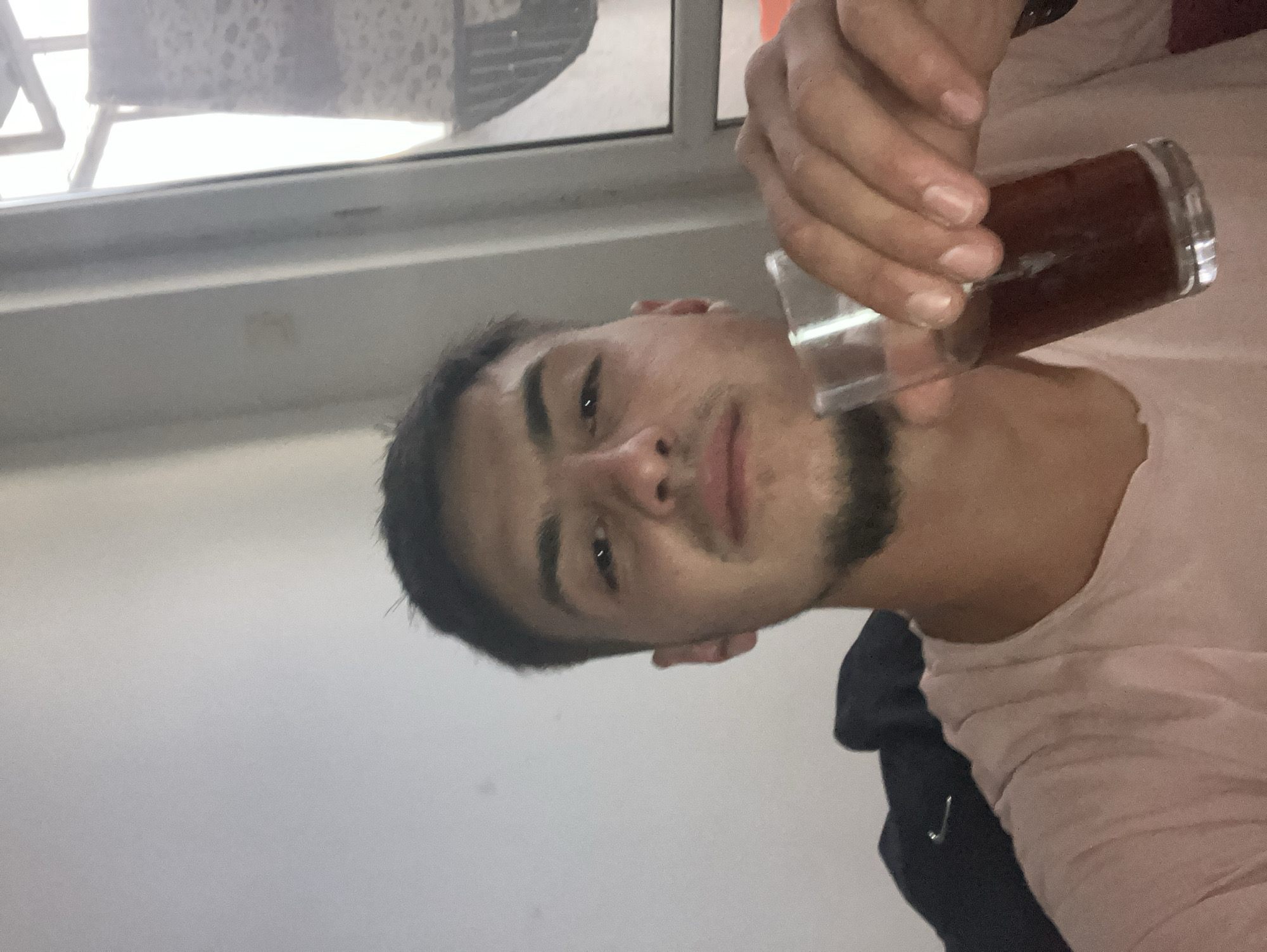 A fair-skinned male with short dark hair and a trimmed beard is holding a glass filled with a dark liquid. He appears to be indoors, near a window with lace curtains. He is wearing a light pink shirt. A dark-colored garment is draped over his shoulder. The location is Plovdiv, Bulgaria.