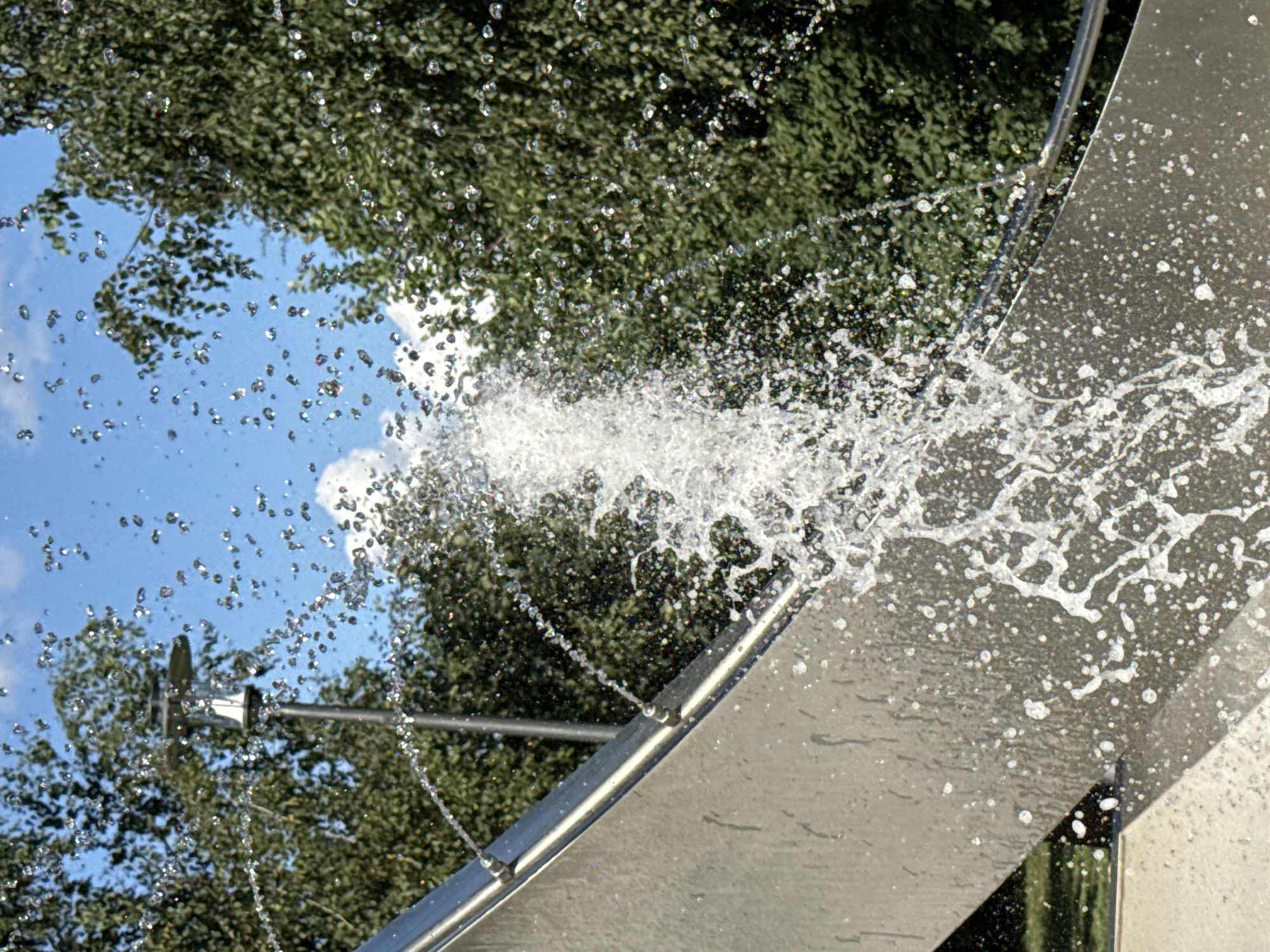 A close-up, slightly low-angle shot captures a modern water feature with a metal structure and a fountain spraying water. The metallic, curved surface of the water feature dominates the foreground, showing water droplets splashing across its textured surface. Behind it, lush green trees form a dense background, their leaves catching the sunlight. In the upper portion of the image, a clear blue sky with scattered white clouds provides a contrast to the greenery. A street lamp with a modern design is partially visible behind the fountain, peeking out from behind the trees. The overall scene suggests an outdoor public space, likely a park or plaza, in the city of Khimki, Russia. There are no people visible in the frame.