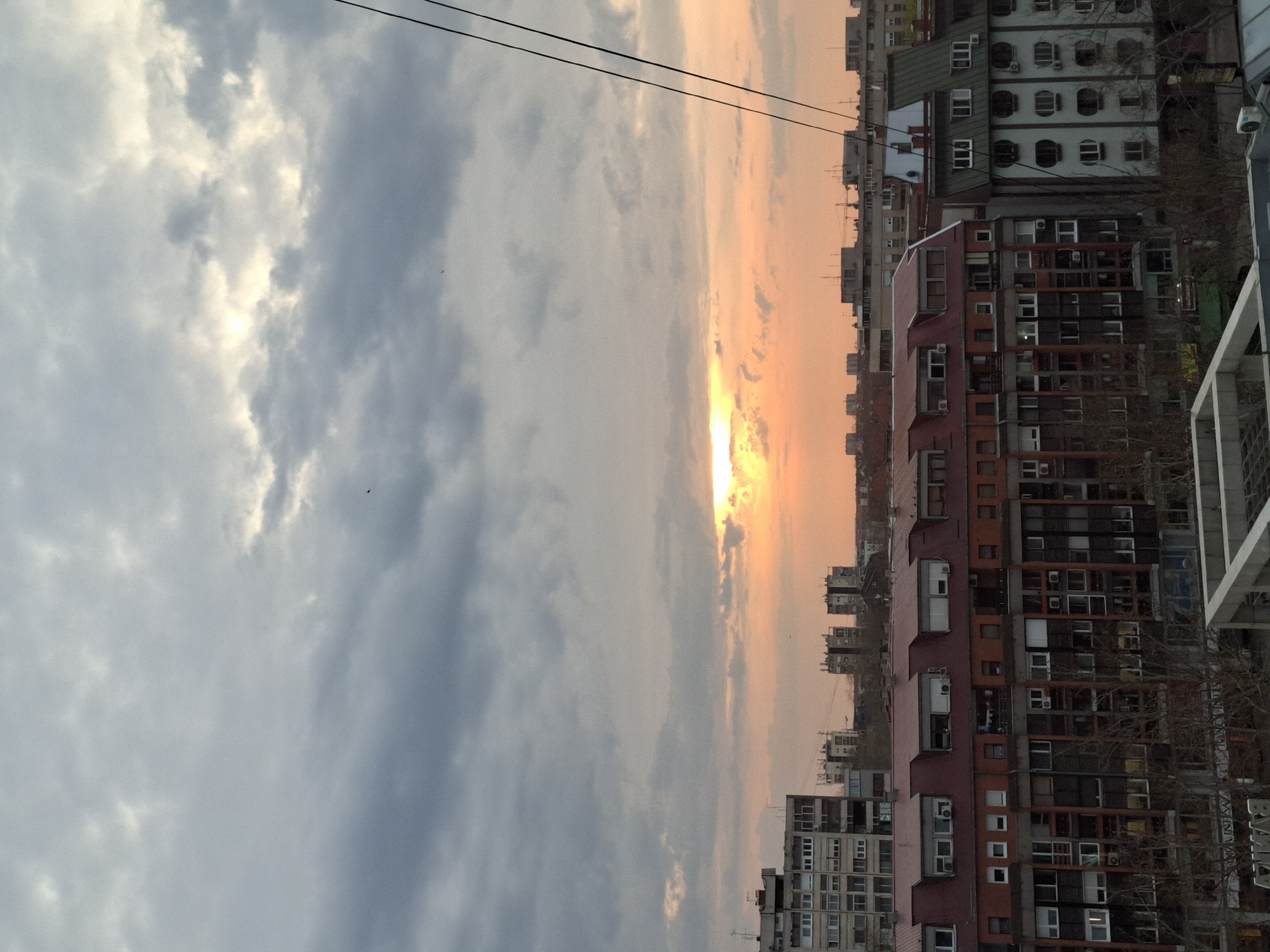 The image shows a cityscape at sunset in Novi Sad, Serbia. Tall residential buildings dominate the right side of the frame, densely packed with windows and varying in color from grey to red. The sky to the left transitions from grey clouds to a bright orange where the sun is setting. Power lines cross the upper portion of the image.