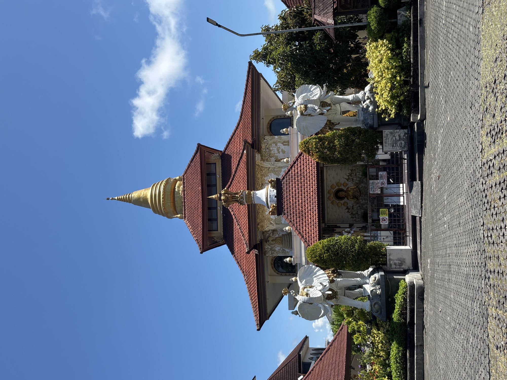 The image shows a Buddhist temple complex in Nusa Dua, Indonesia. The temple has a multi-tiered roof with red tiling and a golden stupa on top. Two white elephant statues flank the entrance. A paved area is visible in front of the temple, with greenery and a street lamp also in view. The sky is blue with some clouds.