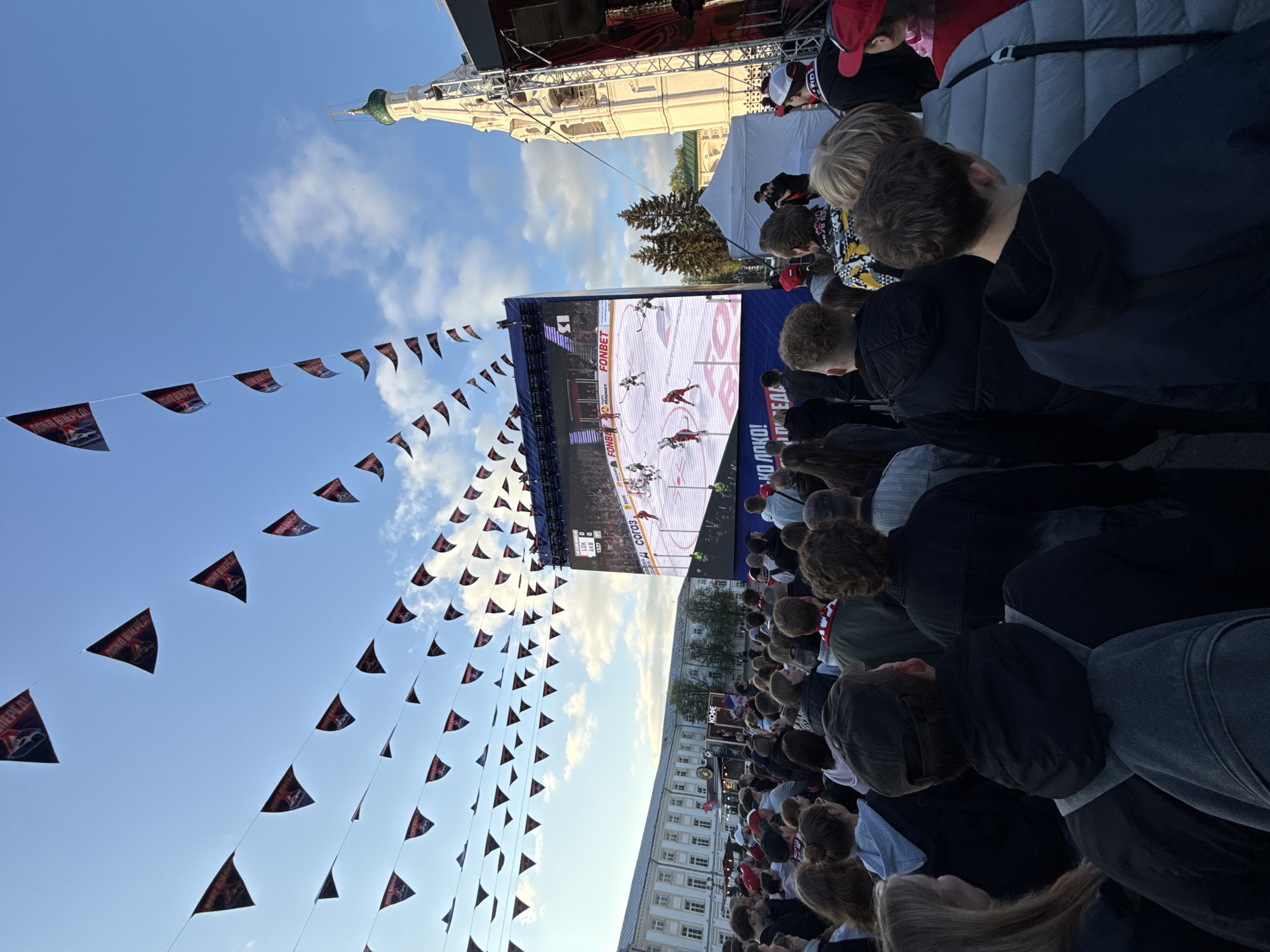 A crowd of people are watching a hockey game on a large outdoor screen. Triangular flags are strung across the top left of the screen. A large building is behind the screen, including its white clock tower. The sky is blue with some clouds. The scoreboard on the screen shows LOK vs AKB. The banner ad says Fonbet. The location appears to be in Заволжье, Russia based on the Russian text.