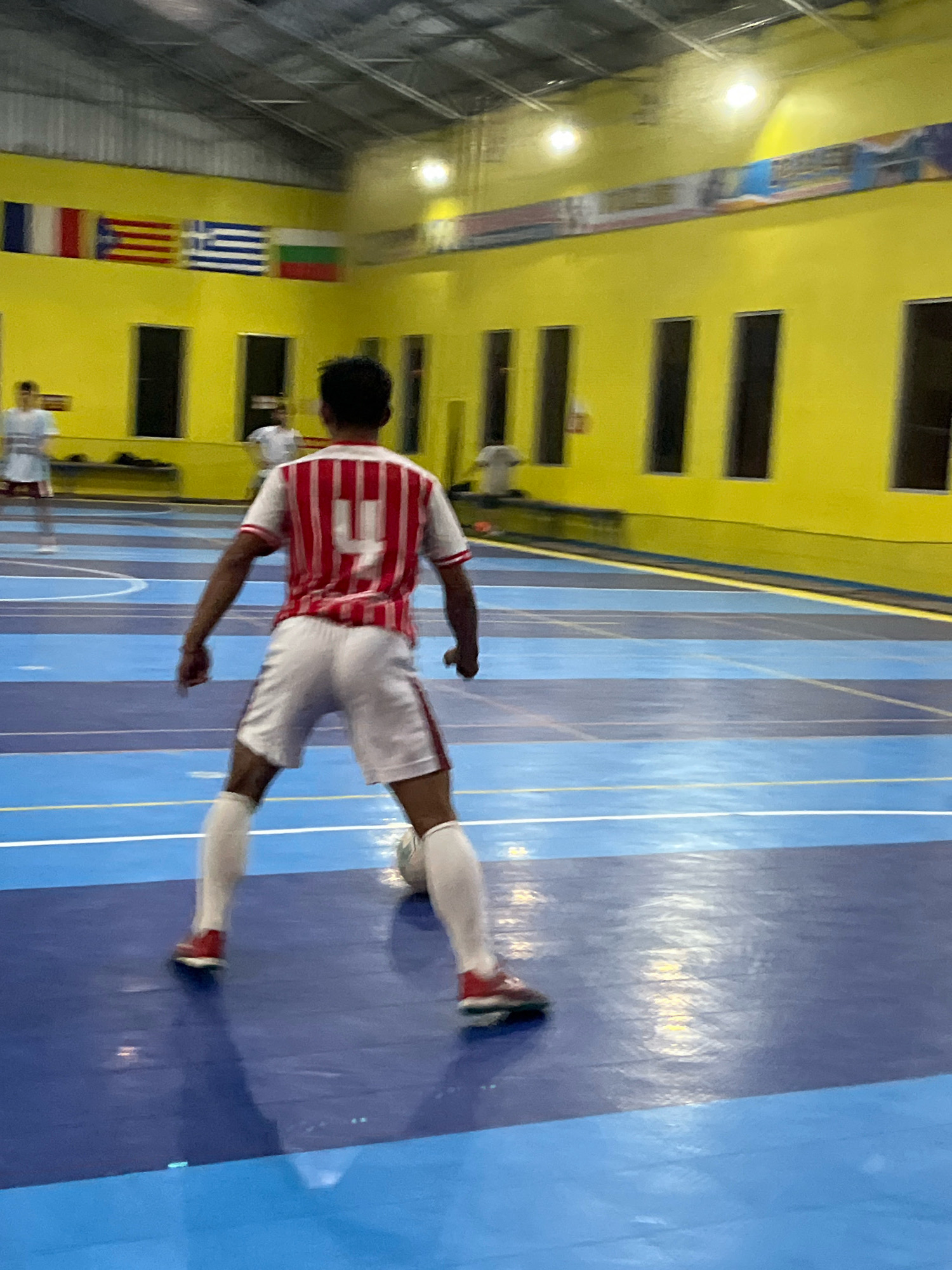 A person is playing soccer in a gymnasium. The person is wearing a red and white striped jersey with the number 4 on the back, white shorts, and white socks with red soccer shoes. The person is kicking a soccer ball on a blue court with blue and white lines. The gymnasium has yellow walls and a silver ceiling with lights. There are flags hanging on the wall, including the French, Spanish, Greek, and Bulgarian flags. There are also several windows on the wall. The image is blurry and taken from behind the person. There are other people in the background, also playing soccer. The image was taken in Jatilawang, Indonesia.