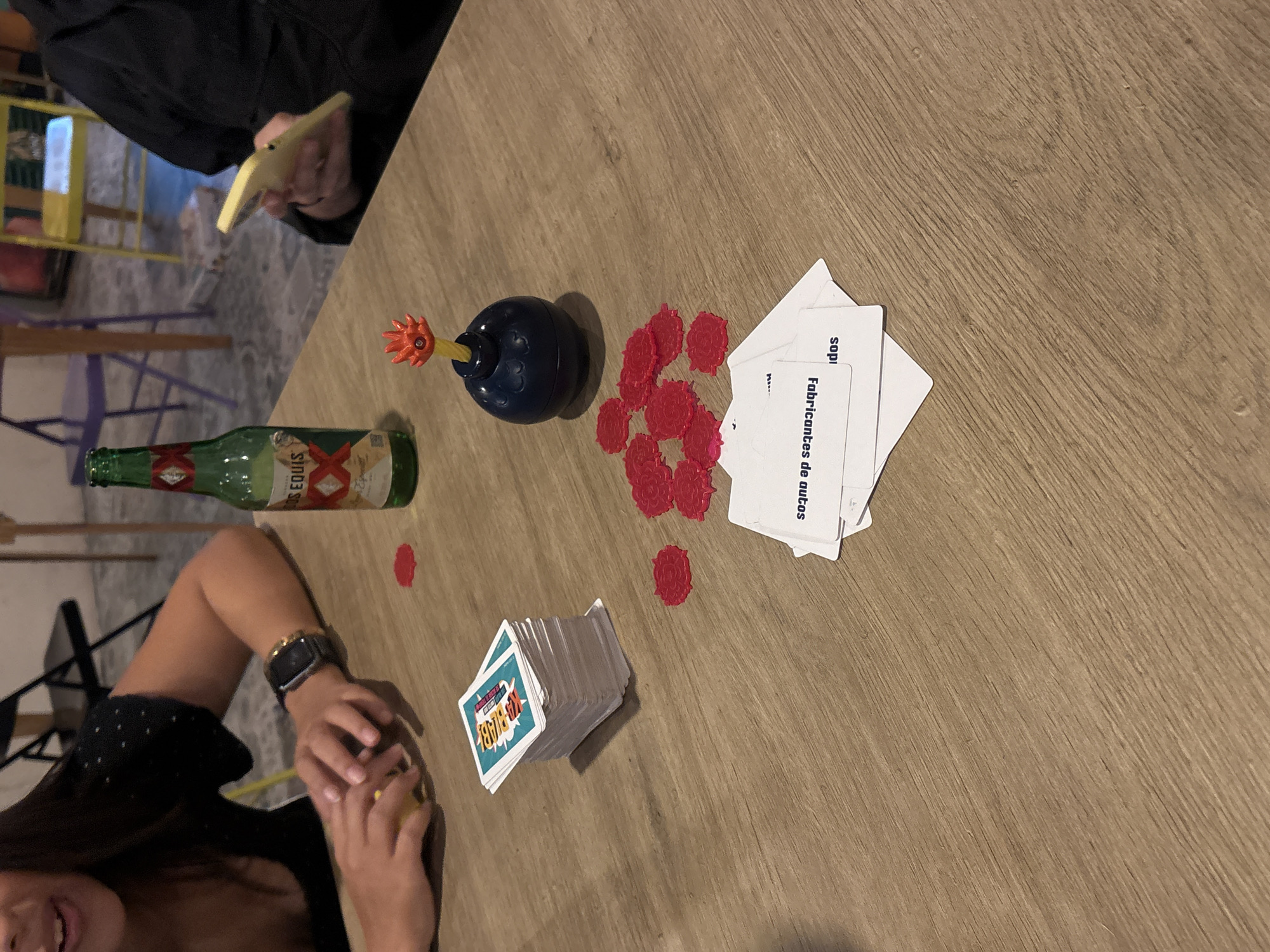 A table in San Andrés Cholula, Mexico, holds a half-empty bottle of XX Equis beer, a toy bomb, several red chips, two stacks of cards, and two people. One person is partially visible and wears a black top and a watch, holding a yellow object. Another person is partially visible and holds a yellow object. One stack of cards has a blue border and is labeled "Kid Babel". Other cards have Spanish text: "Fabricantes de autos" and "Sopi".