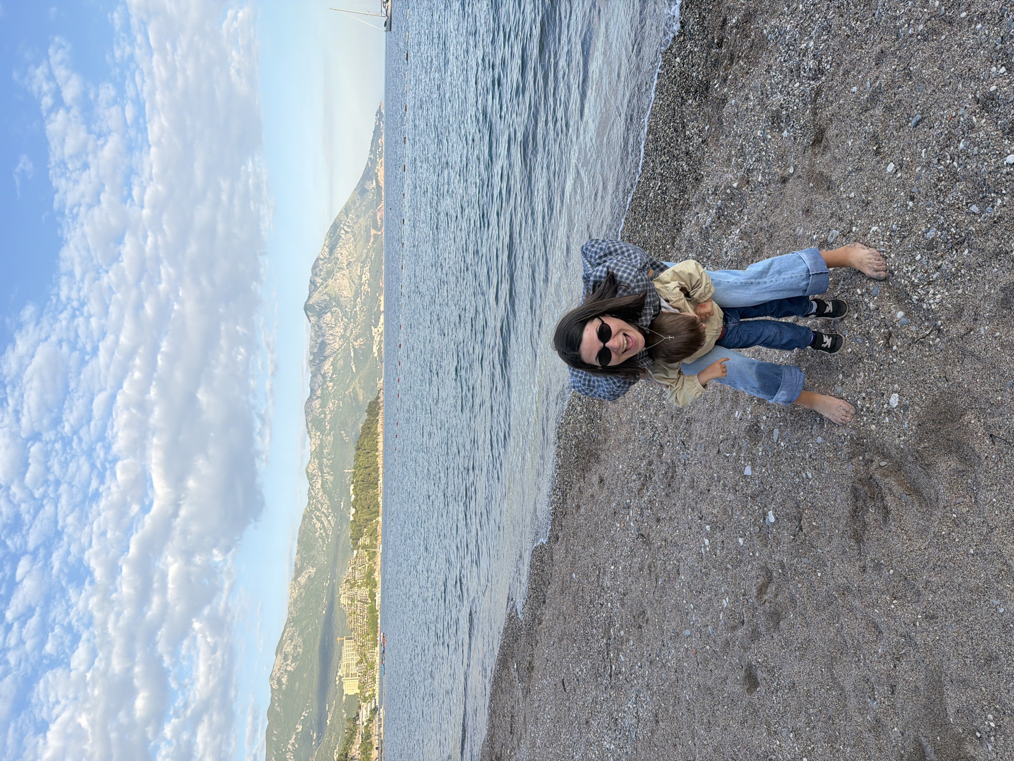 The photograph shows a woman and child on a beach in Budva, Montenegro. The woman is wearing jeans, a plaid shirt, and sunglasses and is holding the child, who is wearing jeans, a jacket, and shoes. They are standing on a pebble beach, and the water is lapping at the shore. In the background is a mountain, the ocean, and a cloudy sky. Buildings can be seen on the edge of the mountain.