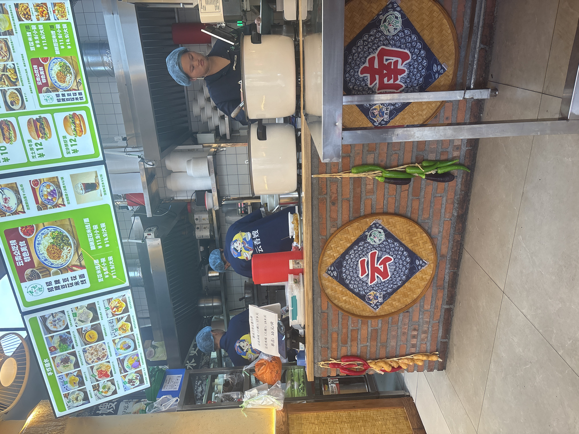 In a casual dining establishment, likely a noodle shop, located in Suzhou Industrial Park, China, the scene depicts a busy kitchen area. Two kitchen staff members, wearing blue hairnets and dark blue uniforms with cartoon graphics, are visible. One staff member is standing at a counter, attending to cooking pots. The counter is made of brick and wood, decorated with large, circular woven mats featuring Chinese characters and imagery. In the foreground, a stainless steel table and a floor tiled in a light grey material are present. Above the counter, a large, illuminated menu board displays various food items, predominantly Chinese dishes, with prices indicated. The overall ambiance suggests a functional and active food service environment.