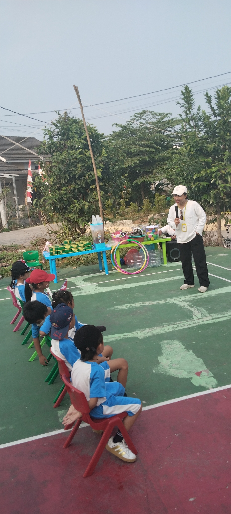 An adult, wearing a white long-sleeved shirt, white cap, and dark pants, stands on a green court holding a microphone and speaking to a group of children. The children are seated on colorful chairs, mostly red and green, facing the adult. They are wearing blue and white uniforms with caps. The scene appears to be outdoors, with trees and vegetation in the background. A table with items like drinks, snacks, and hula hoops is visible behind the adult. A house with a tiled roof and a small Indonesian flag is also in the background. The ground is a combination of green and red surfaces, possibly a sports court. The lighting suggests it is daytime. The overall context suggests an outdoor educational or recreational activity for children.