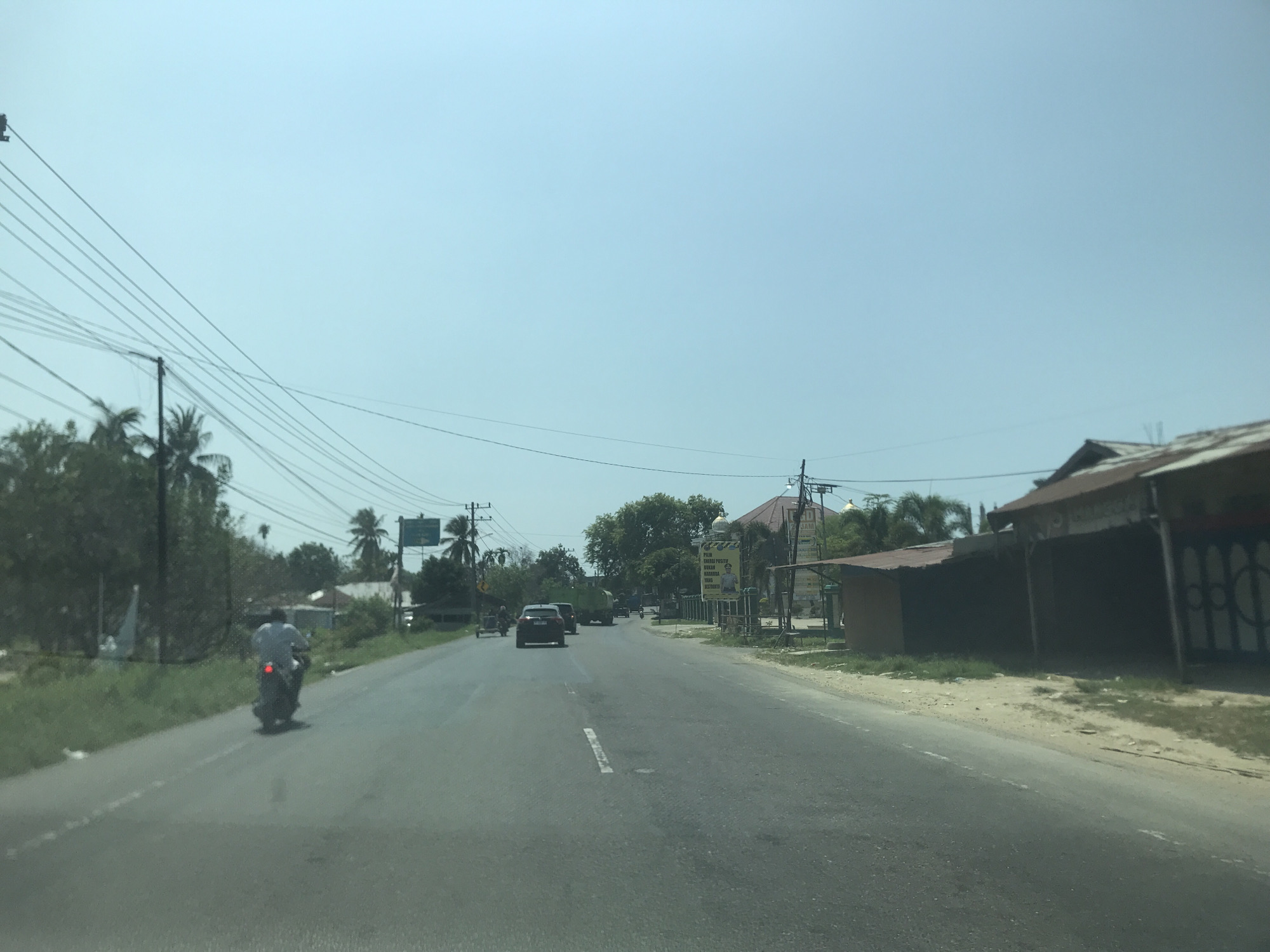 The image depicts a road in what appears to be a suburban or rural area, with a clear sky overhead. On the left side of the road, a person rides a motorcycle. Further ahead, a black car is travelling in the same direction. In the distance, a green truck and another car can be seen. The right side of the road features buildings, one of which appears to be a shop with closed shutters, a gate, and an unpaved area. Utility poles with power lines extend along the left side of the road, and trees are present on both sides. There is an advertisement sign by the right of the road.