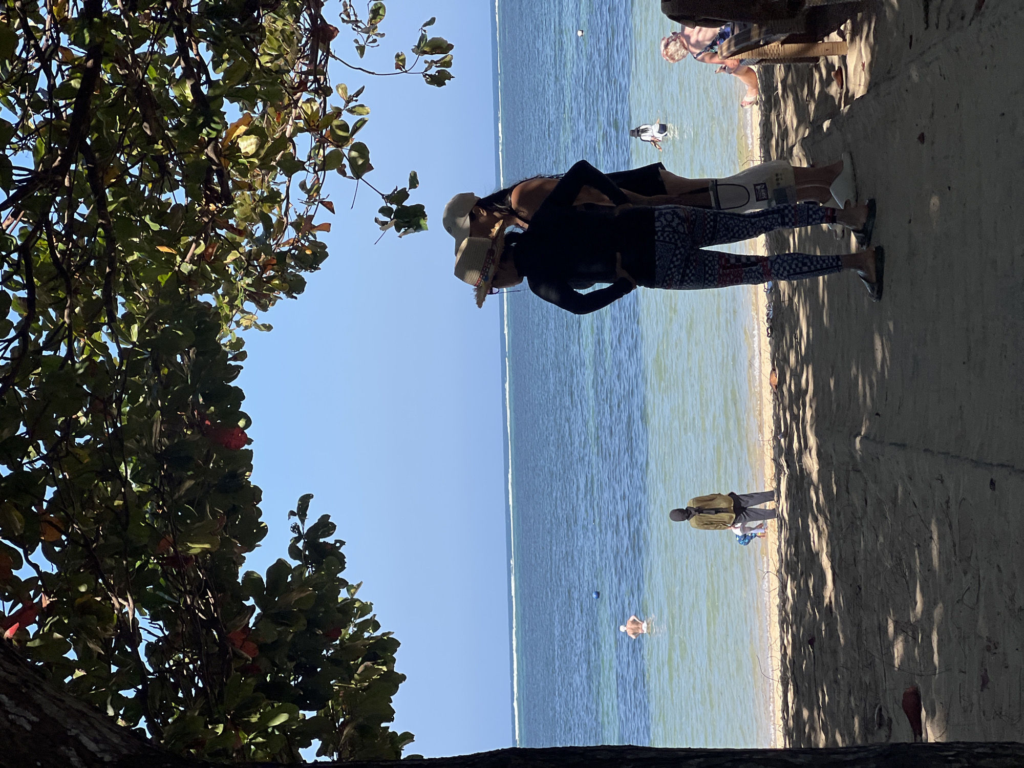 The image depicts a beach scene in Denpasar, Indonesia, on a sunny day with clear blue skies. The foreground is shaded by the branches of a large tree, with its leaves and some red fruits visible. A sandy path leads towards the water. In the middle ground, two women stand facing the ocean. One woman wears a black long-sleeved shirt and patterned leggings, and a straw hat. The other woman wears a black dress and a cap. The ocean is calm, with gentle waves breaking on the shore. Several people are in the water, including a man in the distance and two individuals near the shore. A person dressed in a headscarf and dark clothing stands on the sand near the water's edge, possibly overseeing children playing. In the far right of the image, a person sits on a wooden bench. The overall atmosphere is peaceful and relaxed, indicative of a tropical beach resort.