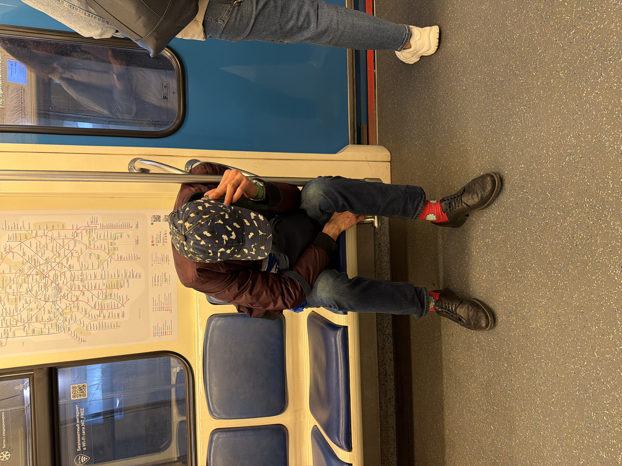 The image shows the interior of a Moscow, Russia subway car. A man wearing a maroon jacket, blue jeans, black shoes with colorful socks, and a patterned cap is seated, seemingly resting his head on the seat. Another person wearing jeans and white sneakers is standing in the background. The interior includes a blue seat, a route map posted on the wall, and advertising stickers on the windows. The subway car floor is textured gray.