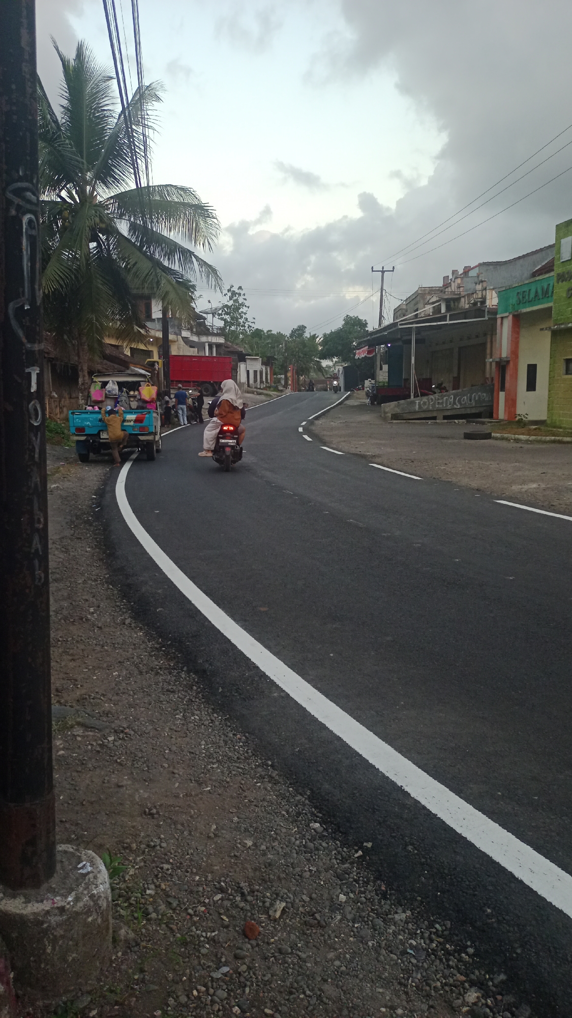 The image depicts a roadside scene in Cogreg, Indonesia, likely during overcast weather. A newly paved asphalt road with a white line marking curves to the right. On the left side of the road, a rough, gravelly shoulder is visible, bordered by a concrete post with writing on it. A coconut palm tree stands tall on the far left.
In the mid-ground, a light blue pickup truck is parked, its bed filled with various bags. A person, possibly a child, is climbing onto the truck. A group of people are standing near the truck. Further down the road, a motorcycle carrying two passengers, both wearing headscarves, is traveling away from the camera. The rider wears an orange headscarf and a beige hijab, while the passenger wears a beige headscarf and a brown outer garment.
The right side of the road features a row of buildings, including a structure with a green facade and a sign that reads "SELAMAT". Other buildings appear to be shops or small businesses. A red truck is parked in the distance, and more buildings line the street. The sky is filled with dark, heavy clouds. The overall impression is of a semi-urban or peri-urban area in Indonesia.