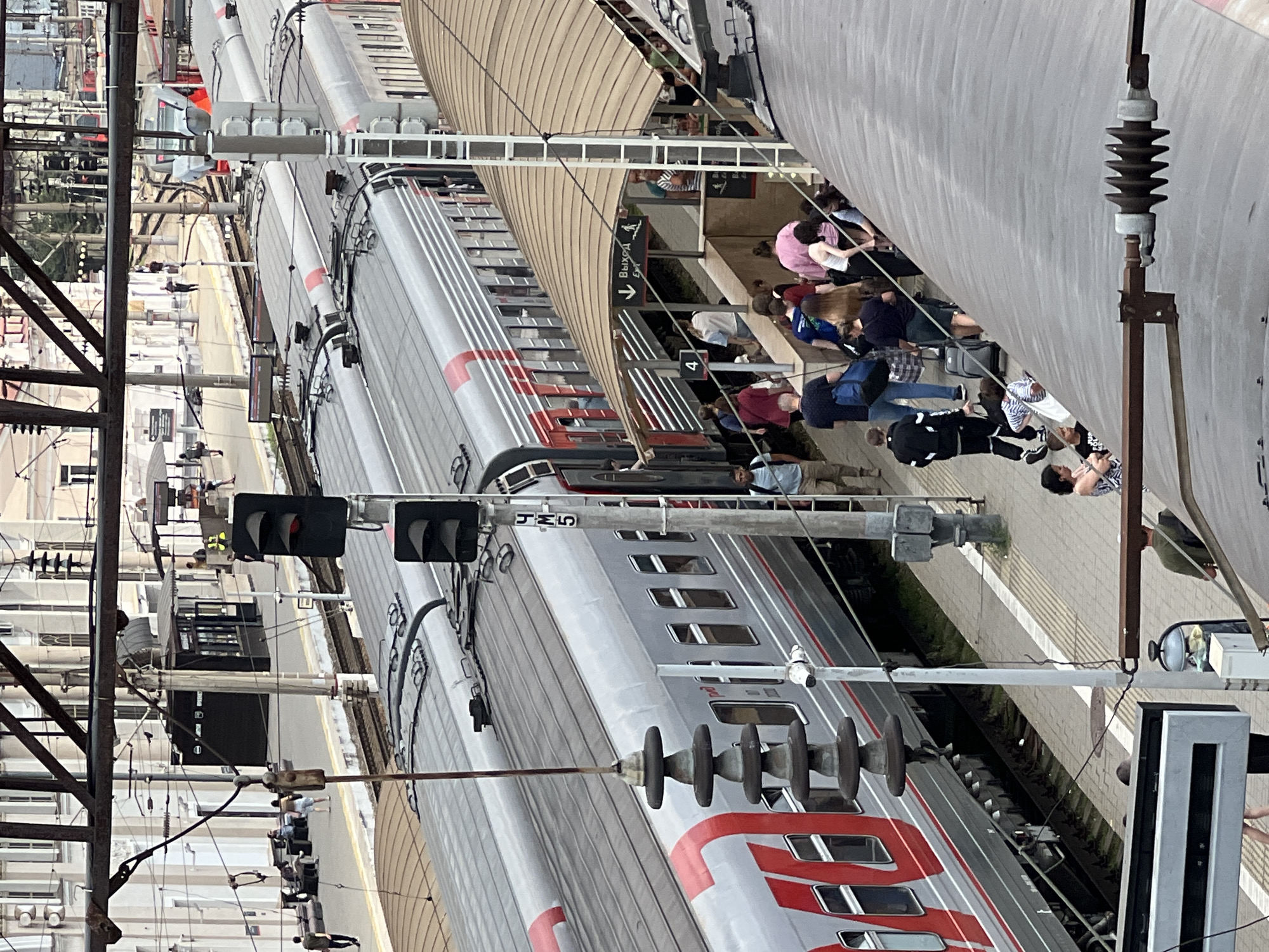 The image shows a train station platform in Krasnodar, Russia, with two trains parked side-by-side on adjacent tracks. Several people are visible on the platform, some waiting to board the trains, while others are disembarking. A covered walkway with an "Exit" sign is visible, leading away from the platform. The trains are modern passenger trains, with silver and red livery. Overhead power lines and signal lights are present. The platform itself is paved and has yellow tactile paving for the visually impaired. The architecture of the station buildings is visible in the background, featuring classical styling with light-colored facades. The overall scene suggests a busy transportation hub during the day.
