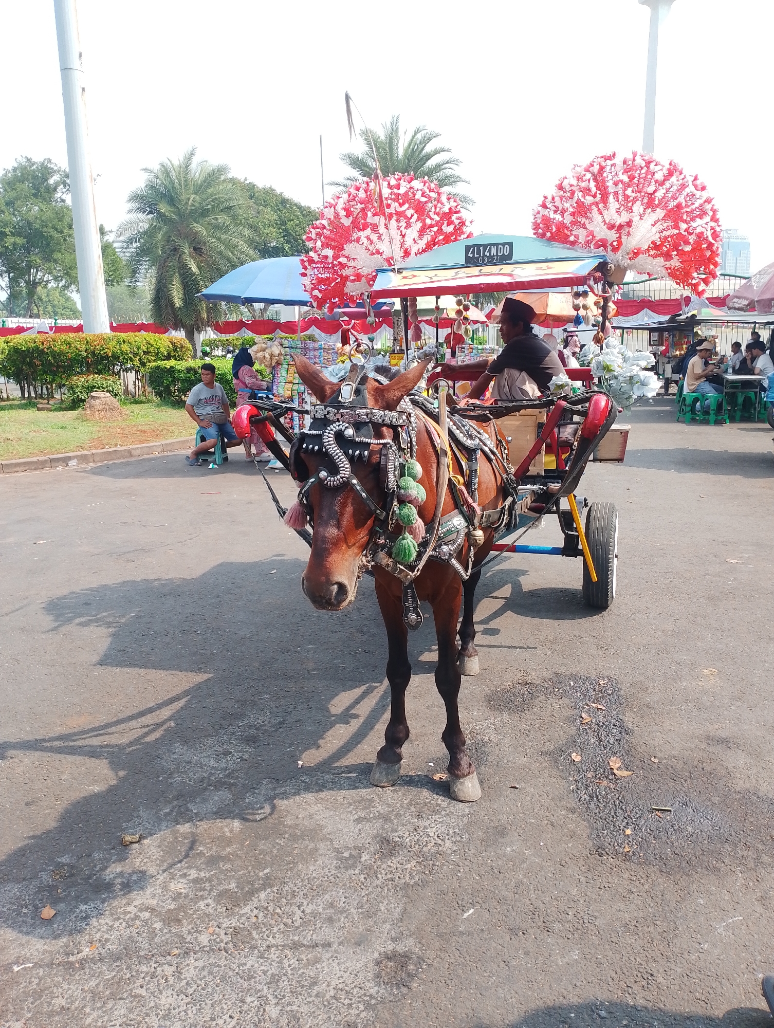 In a paved outdoor area, a brown horse is harnessed to a small, two-wheeled cart. The horse faces the camera, and its head is adorned with decorative tack. The cart appears to be a mode of transport or a mobile stall, with a canopy overhead decorated with red and white floral-like ornaments. A person wearing a dark shirt and a peci cap is seated on the cart, appearing to be the driver or operator. In the background, several people are seated at tables, some eating and others looking at their phones, suggesting a public space with amenities. Beyond them, there are trees, greenery, and structures that suggest an urban park or recreational area. A tall, white pole, possibly a monument or utility pole, is visible in the far background. The overall scene suggests a lively outdoor setting in a city. The inscription "4L14NDO" is visible on a sign on the cart's canopy. This scene is likely located in the National Monument (Monas) park area in Central Jakarta, Indonesia, a popular tourist destination known for its open spaces and occasional recreational activities like horse-drawn carts.