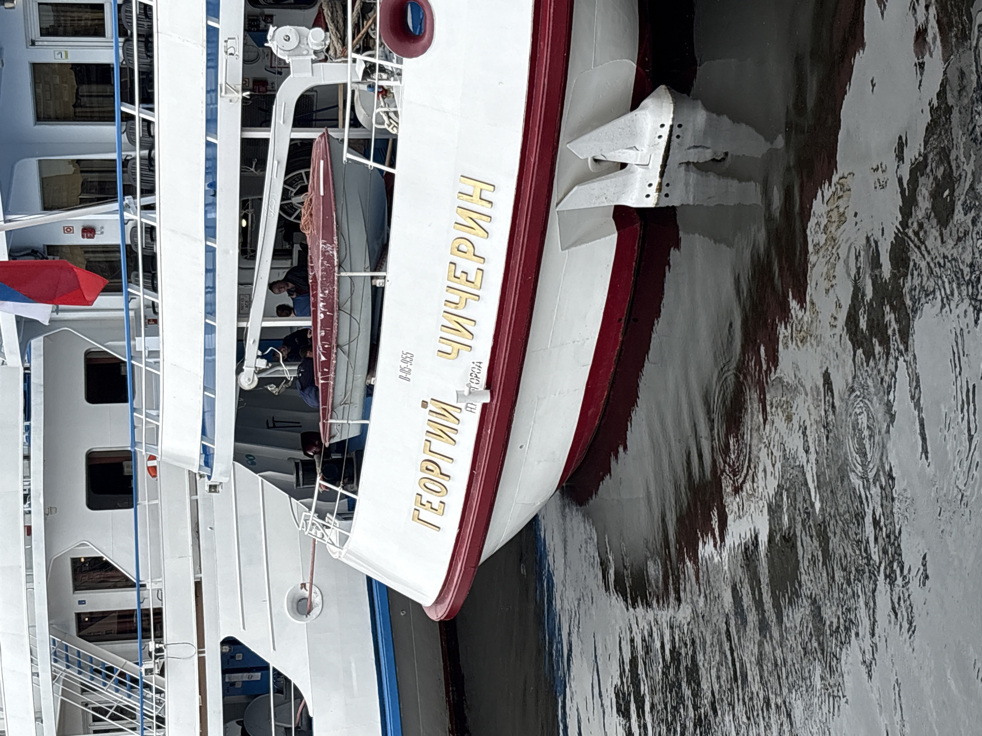 A close-up, eye-level shot captures the stern of a white passenger ship named "ГЕОРГИЙ ЧИЧЕРИН" (Georgiy Chicherin) docked in the city of Shushary, Russia. The ship's name is prominently displayed in gold lettering on its white hull, which is accented by a deep red stripe along the waterline. Two white propulsion pods are visible at the stern, partially submerged in the dark, rippling water. The water reflects the ship's red hull and white superstructure. In the background, another white ship is partially visible. A small red life raft is secured on the deck of the Georgiy Chicherin, and several people are seated in the background, near the life raft, appearing to be passengers. A Russian flag, predominantly red and white with a blue stripe, flies from a mast on the ship. The scene suggests a calm day at the harbor.