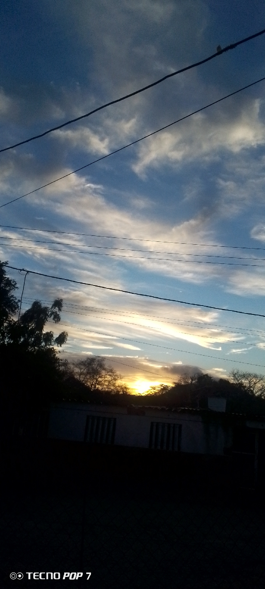The image shows a sunrise or sunset in Altagracia, Venezuela. The sky is blue with white clouds and several power lines are visible in the foreground. A building with dark windows and trees are silhouetted against the horizon where the sun is setting or rising. The lower part of the image is dark. A phone brand watermark is present on the bottom left corner.
