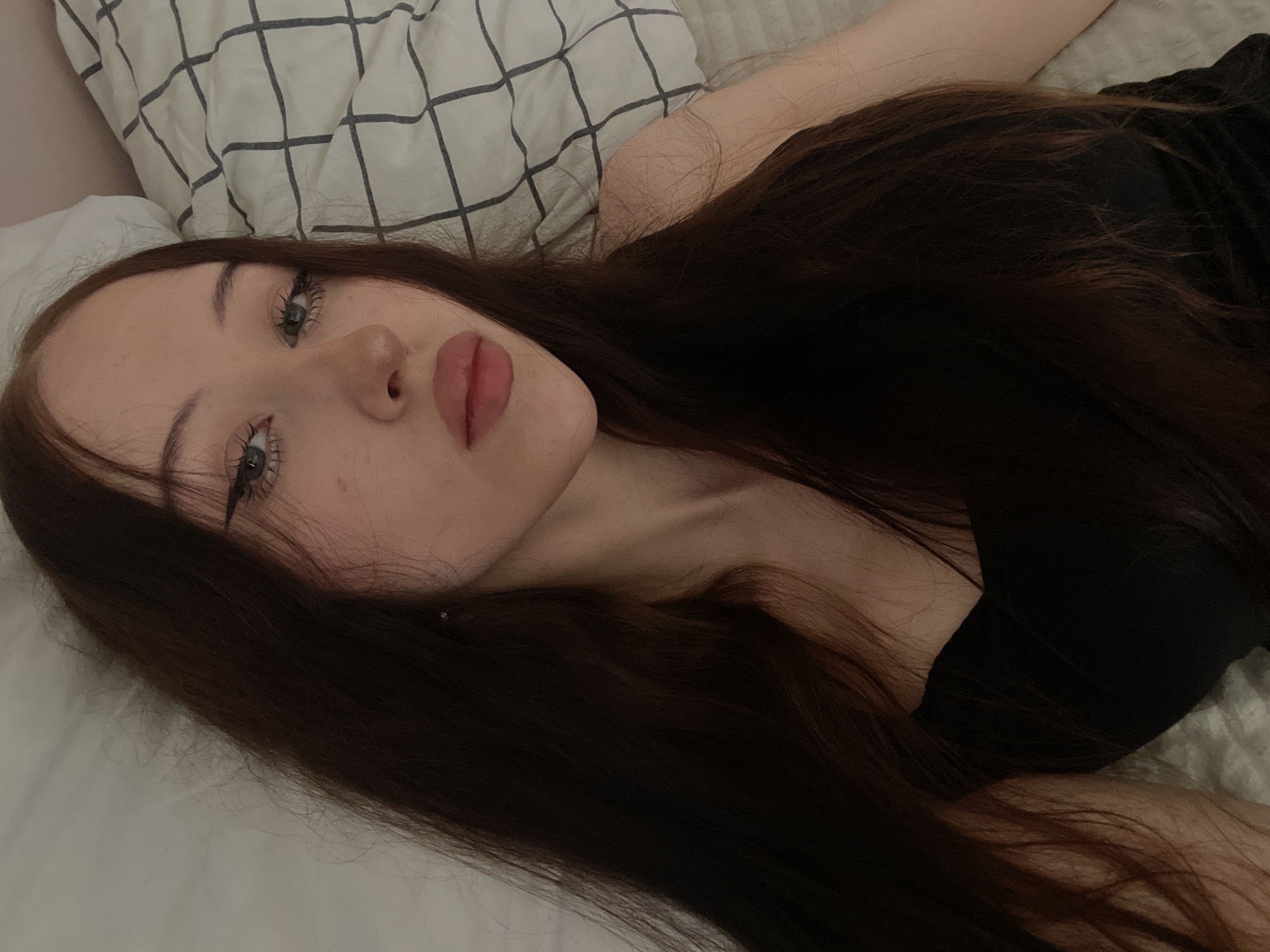 A young woman with long, dark brown hair lies on a bed. She is wearing a black top. Her head is tilted to the side, and her eyes are open, looking towards the camera. She has on winged eyeliner and lip gloss. The background shows a white pillow with a grey grid pattern. The scene appears to be indoors, likely a bedroom. There is no information in the image to determine the location in the city of Votkinsk, Russia.