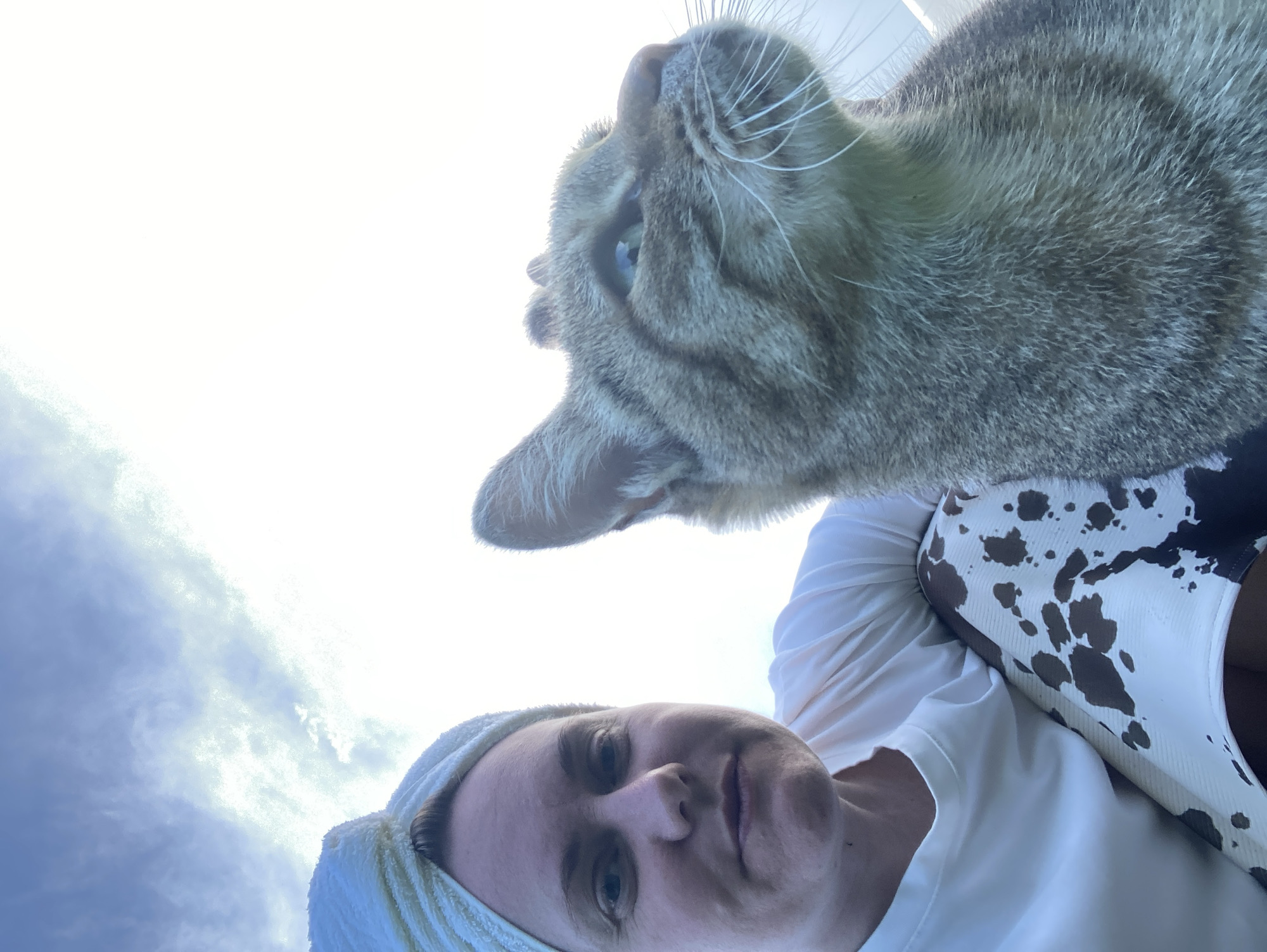 In an outdoor setting, a woman and a cat are photographed from a low angle against a sky with light clouds. The woman, positioned on the left of the frame, wears a white t-shirt and a white towel wrapped around her head. She is looking forward with a neutral expression. To her right, a tabby cat is visible in close-up, with its head tilted upwards and to the right, looking off-camera. The cat has distinct tabby markings and its fur appears textured. The woman is wearing patterned pants with brown and white splotches. The background consists of a bright sky, suggesting it is daytime, possibly late afternoon or early morning due to the soft lighting. The specific location in the city of Bolshoy Bukor, Russia, is not identifiable from the visual information provided.