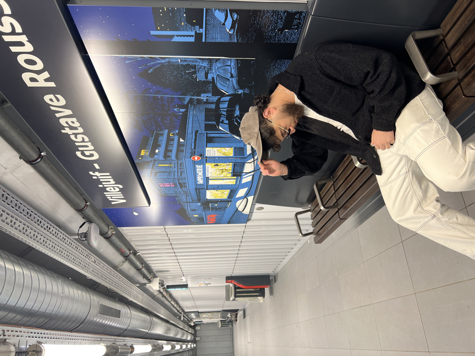 The image depicts a male subject seated on a wooden slatted bench in a public transport station. The location is identified by an illuminated overhead sign reading "Villejuif - Gustave Roussy," indicating a metro station in Villejuif, France. The man wears a dark, textured jacket over a white shirt, light-colored pants with visible contrast stitching, sunglasses, and a grey baseball cap, which he is adjusting with his right hand while looking downwards. Behind him is a large wall mural illustrating a stylized nighttime urban street scene with buildings, an "IMPRIMERIE" (printing shop) sign, and vehicles, rendered in shades of blue with yellow illuminated windows. The station environment features a light grey tiled floor, white vertical corrugated wall panels, and an exposed ceiling infrastructure with metallic ventilation ducts, electrical conduits, and fluorescent strip lighting. A dome security camera and a white wireless access point are mounted overhead. Further down the corridor to the left, a red and black ticketing machine and a grey waste bin are visible against the white wall.