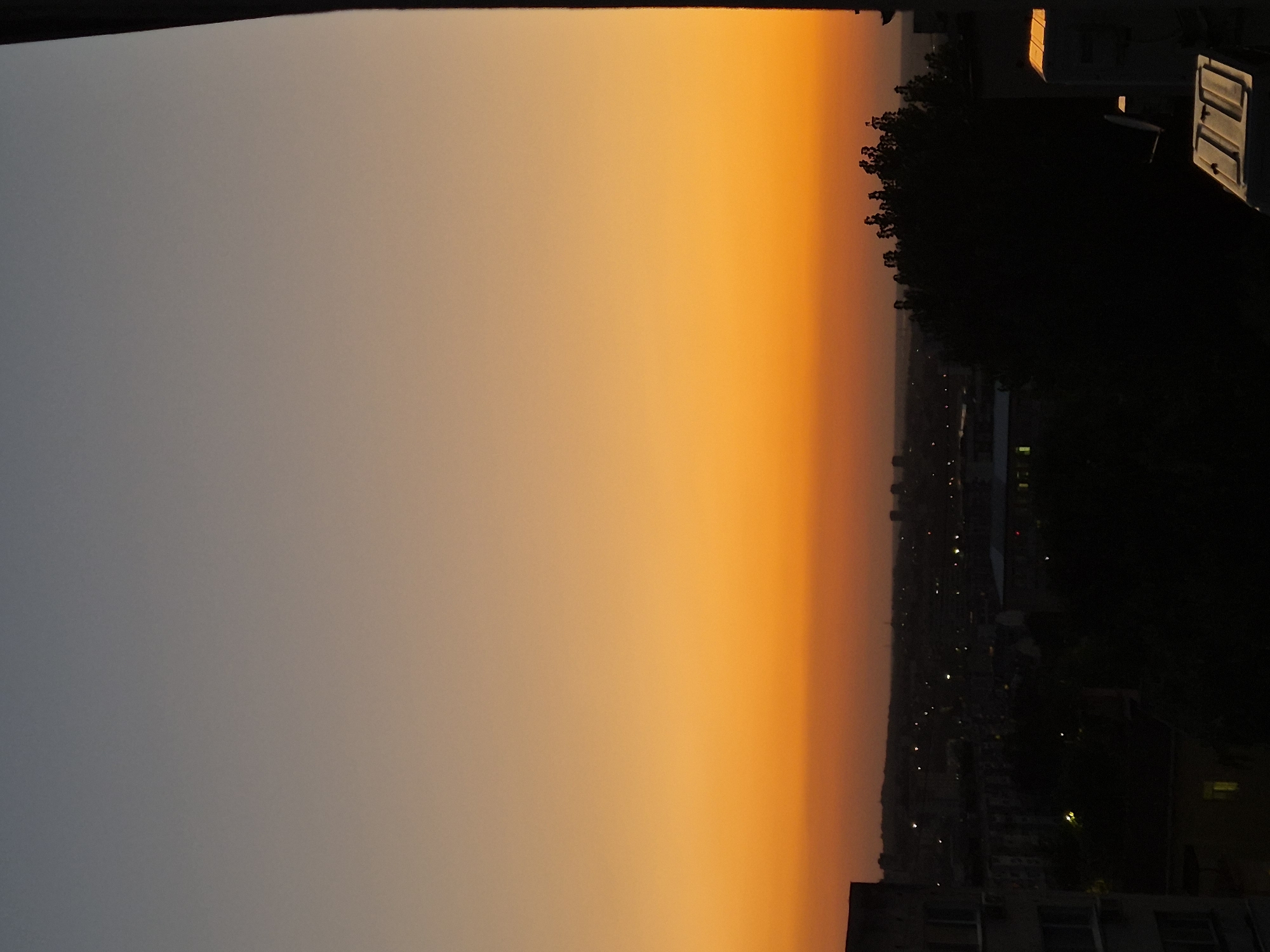 The image depicts a dusky cityscape at sunset, with a gradient of colors in the sky transitioning from a muted gray at the top to a vibrant orange near the horizon. In the foreground, the silhouettes of buildings and trees are visible against the diminishing light. The city is partially illuminated by scattered lights, suggesting it is an urban environment. On the right edge of the frame, a dark vertical element, possibly a building facade or a structural beam, partially obscures the view. The overall scene conveys a sense of twilight in a city.