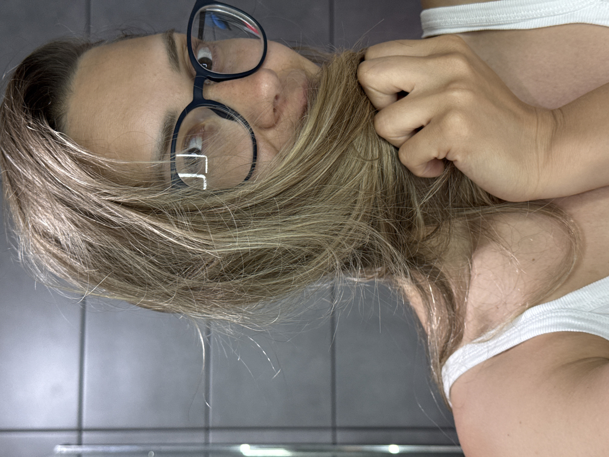 A young woman with blonde hair, wearing glasses and a white tank top, is captured in a close-up, self-taken photograph against a gray tiled wall. Her hair is partially obscuring her face as she holds a section of it with her right hand. The image is a selfie taken from the chest up, focusing on her upper body and face. The lighting appears to be natural, casting subtle shadows. The background is a wall of rectangular gray tiles, suggesting an indoor setting such as a bathroom or shower. There is no indication of the city of Chaikovsky, Russia.