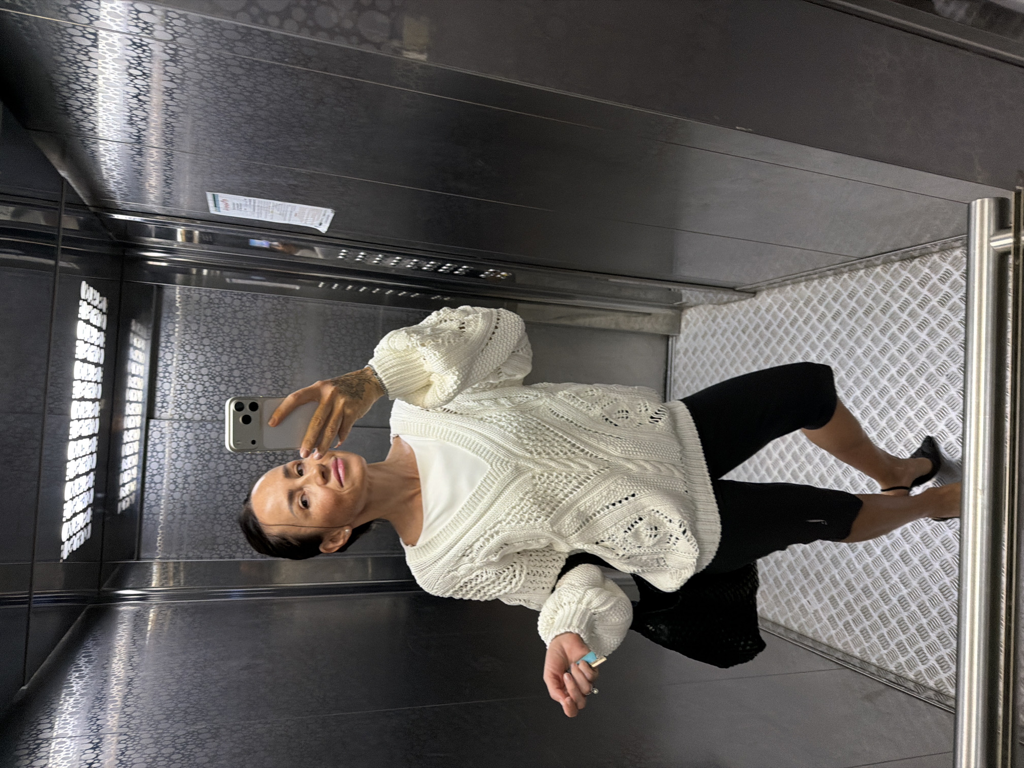 A woman is taking a mirror selfie in an elevator. She is wearing a white cable-knit sweater with a white undershirt, black cropped leggings, and black heeled shoes. She has a black bag on her left arm. The elevator has silver metallic walls with a circular pattern and a diamond-plate floor. The image is taken from the waist up, focusing on the woman and her reflection. The location is the city of Khimki, Russia.