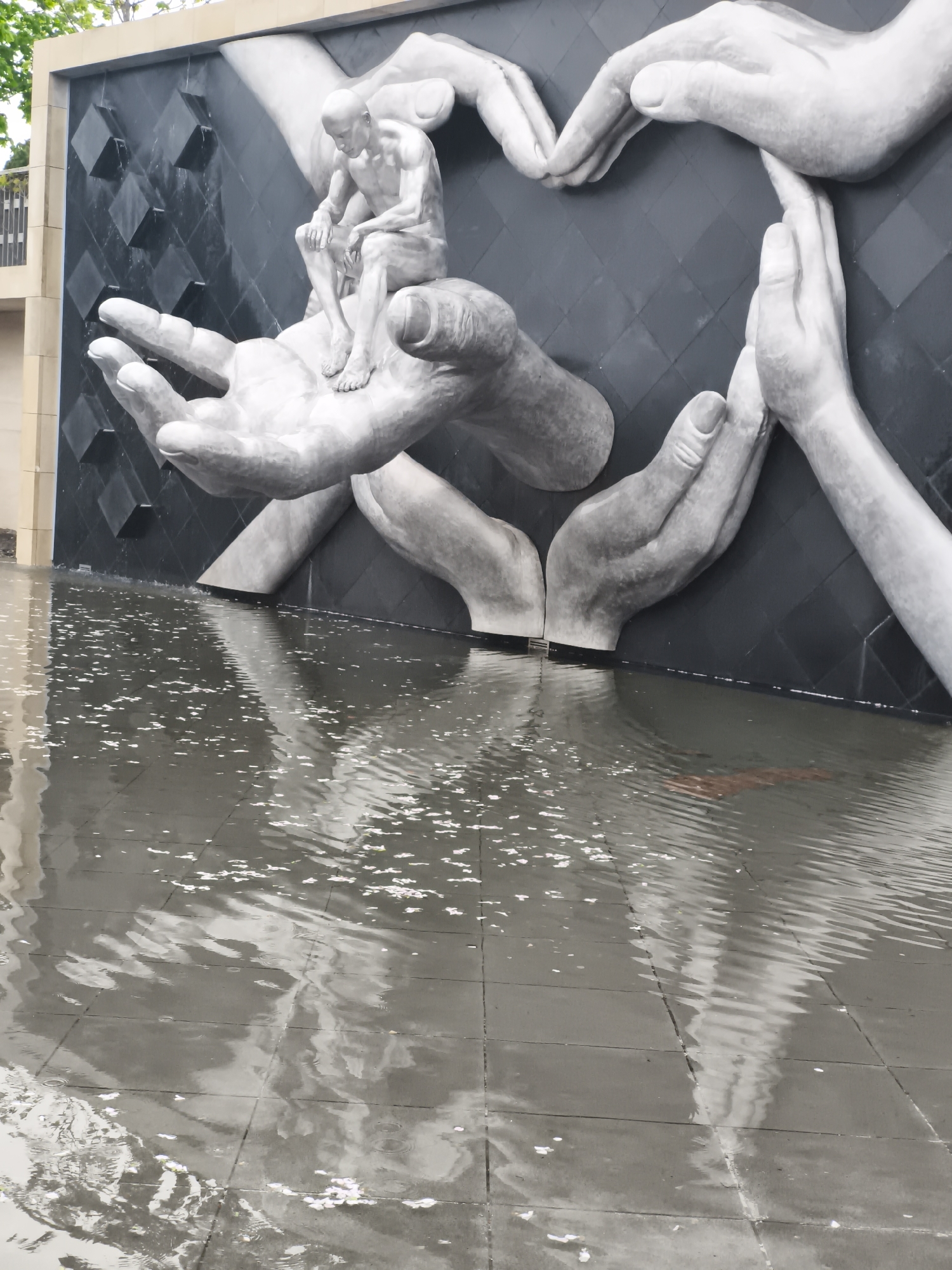 The image depicts a sculpture located in Krasnodar, Russia. The sculpture features a nude male figure seated in the palm of a large, sculpted hand. Several other hands frame the scene, forming an abstract, supporting structure. The backdrop behind the hands is a dark, textured surface. The sculpture appears to be partially submerged in water, with the tiled ground around it also covered in water. Small white objects, potentially petals or debris, float on the water's surface.