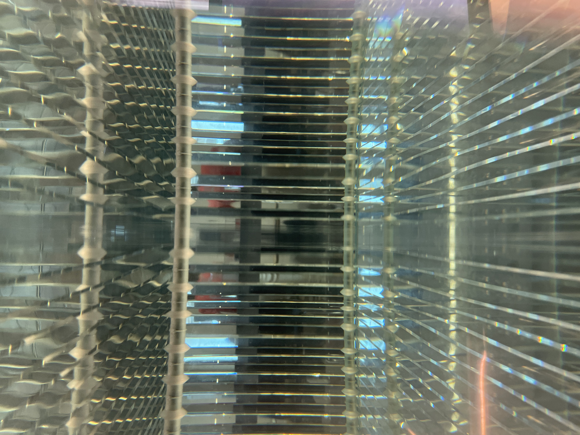 The image presents an abstract, refracted view through a clear, structured object with repeating horizontal and corrugated elements. The horizontal elements appear as transparent shelves or plates, stacked with narrow gaps in between. These are seemingly supported by vertical structures marked with regularly spaced circular or cylindrical features. Refraction and internal reflection within the object create a complex network of lines and shapes, distorting the view of what lies beyond. Hints of color, especially blues and reds, appear within the refractions, suggesting a light source or objects outside the frame. No people are visible. The origin or significance of the photograph in the city of Belgrade, Serbia cannot be confirmed.