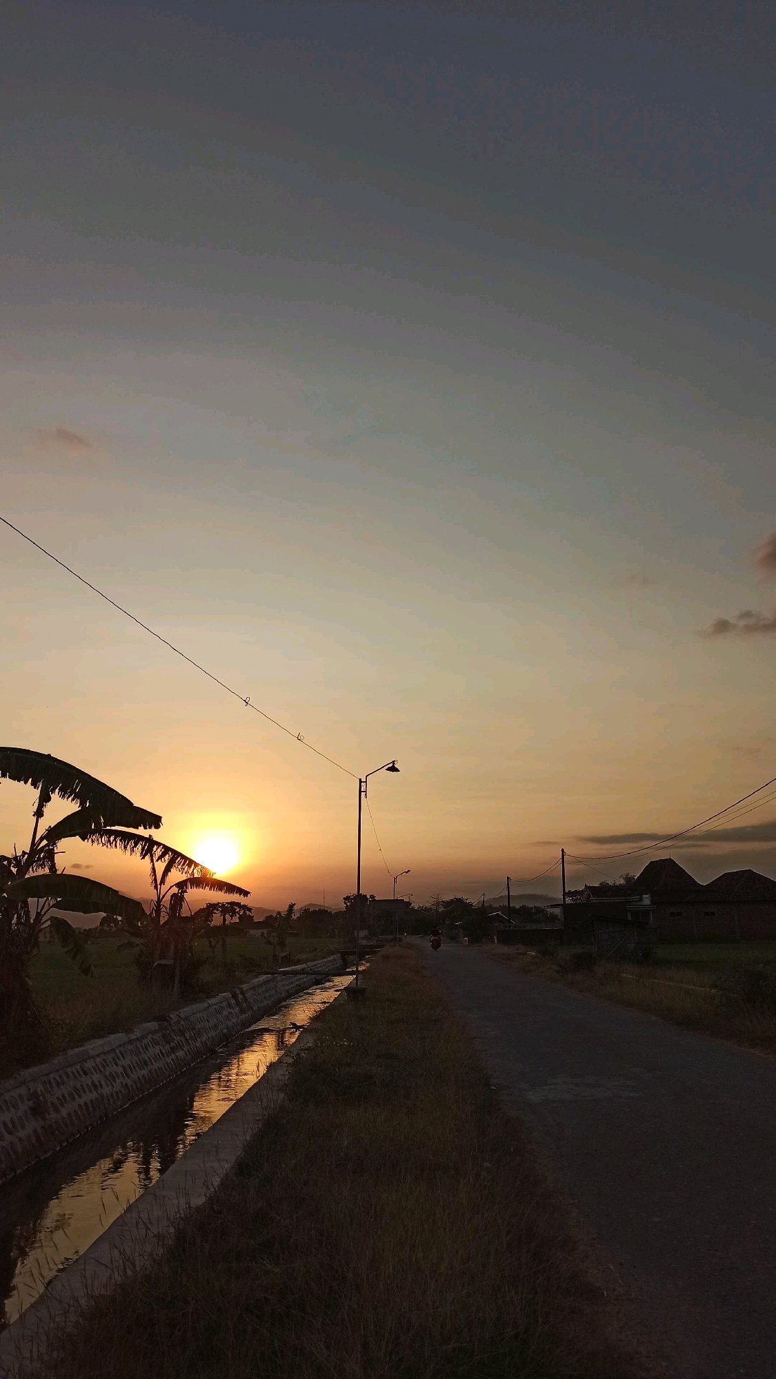 The image depicts a rural roadside scene at sunset in Josari, Indonesia. A paved road runs diagonally from the lower right to the upper center of the frame, flanked by a grassy embankment on the right and a concrete-lined irrigation ditch filled with water on the left. The water reflects the warm hues of the setting sun, which is visible on the left side of the image, peeking through the fronds of banana trees. A utility pole with a street lamp stands to the right of the road. In the distance, several houses with traditional roof structures are visible. A single motorcycle with a rider is moving away from the viewer on the road. The sky transitions from a pale blue at the top to a warm orange and yellow near the horizon, with a few wispy clouds. The overall atmosphere is serene and characteristic of a village setting at dusk.