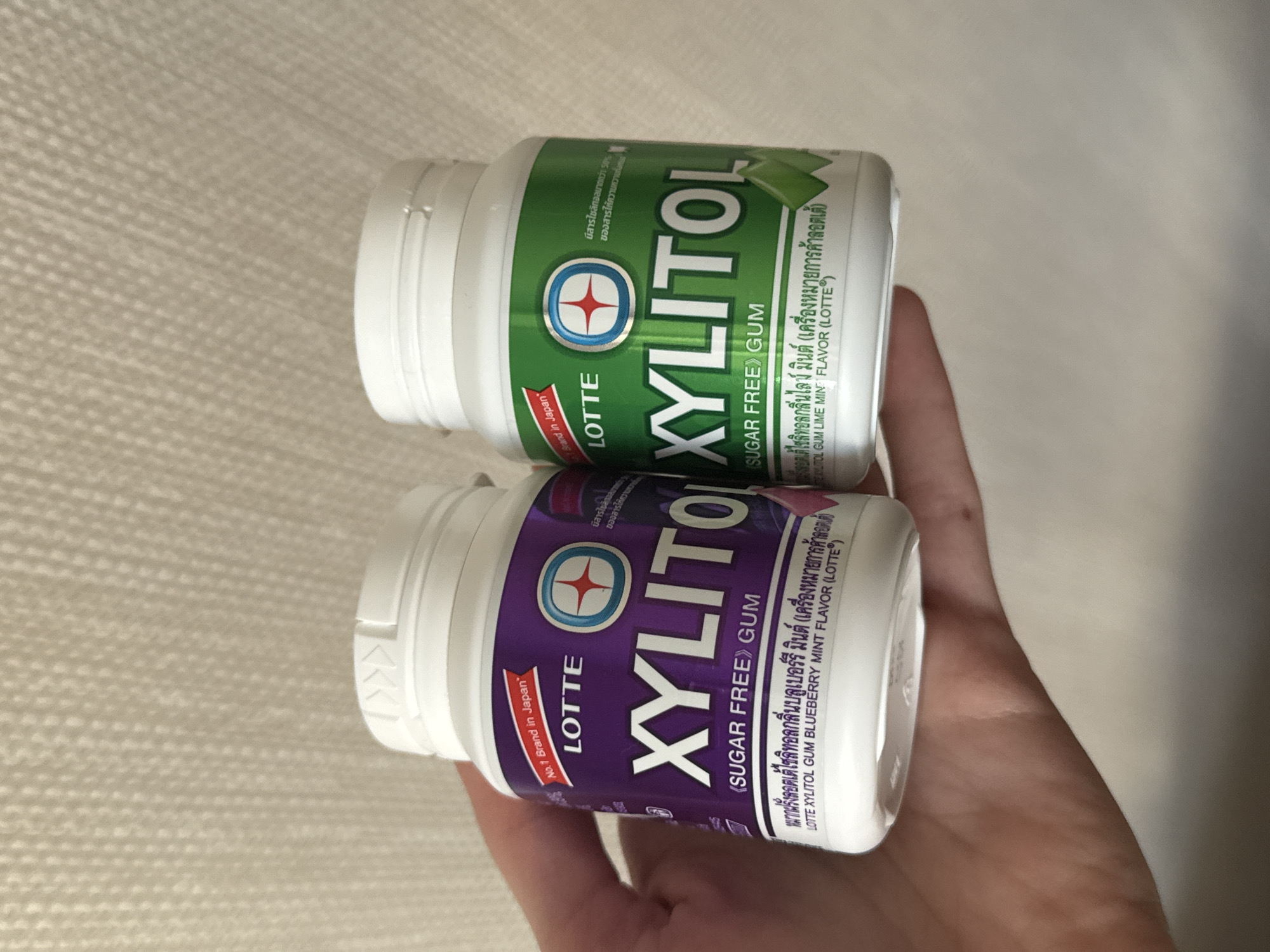 An indoor, close-up shot captures a hand holding two containers of Lotte Xylitol chewing gum, presented slightly askew. The background consists of a light-colored, textured surface, possibly wallpaper or fabric.
The subjects are a human hand and two distinct flavors of Lotte Xylitol gum. One container is predominantly green, while the other is purple. Both are cylindrical, made of plastic, and feature white screw-on caps.
The activity shows a person displaying or holding the gum containers.
Notable details include the branding and product information visible on the labels. Both containers feature the "LOTTE" logo and "XYLITOL" in large lettering, with "SUGAR FREE GUM" directly underneath. The purple container clearly states "No. 1 Brand in Japan" on a red banner. Both labels include text in Thai and English.
On the green container, visible Thai text translates to "Contains more than 50% Xylitol of all sweeteners" and "LOTTE XYLITOL GUM LIME MINT (LOTTE® trademark)," followed by "LOTTE XYLITOL GUM LIME MINT FLAVOR (LOTTE®)."
On the purple container, similar Thai text is visible, translating to "LOTTE XYLITOL GUM BLUEBERRY MINT (LOTTE® trademark)," followed by "LOTTE XYLITOL GUM BLUEBERRY MINT FLAVOR (LOTTE®)." Part of the "Contains more than 50% Xylitol..." Thai text is also visible on the purple container.