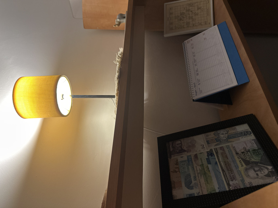 An interior scene within a building in Plovdiv, Bulgaria, displays two wooden shelves against a light-colored wall, illuminated by a warm light source. On the upper shelf, an illuminated table lamp with a pleated, cylindrical, yellow-orange fabric shade rests on a dark metal stand. To its left, a tangled bundle of light-colored string or yarn is visible. Further to the right on this shelf, a small grey elephant figurine with white tusks stands on a white rectangular base. Below, on a second wooden shelf, from left to right, are three items: a black rectangular picture frame containing a collage of various international banknotes, including visible text such as "100," "Canada," "Bank of Jamaica," and "Banco Central del Paraguay." Next to it, a white coil-bound desk calendar with a blue base is open to "Март - March," displaying a daily schedule grid for approximately March 9-13. To the right of the calendar, a smaller rectangular picture frame with a white border contains a sepia-toned image resembling an old map or schematic diagram. The overall lighting is dim, with the lamp casting direct illumination and highlighting the objects.