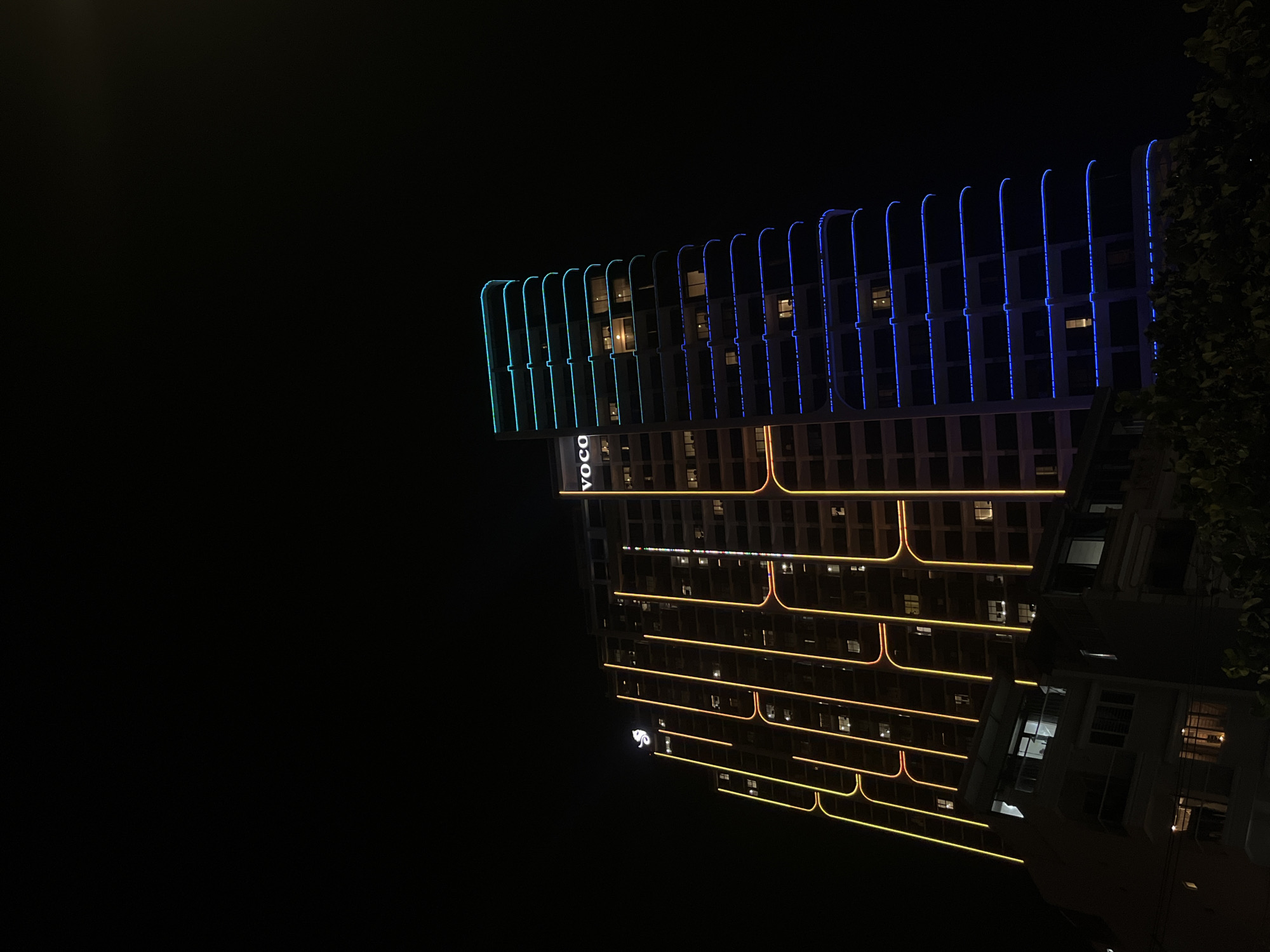The image displays a modern building at night in Nha Trang, Vietnam. The building's facade is illuminated by vibrant neon lights. The top of the building features a series of curved, vertical lights in alternating turquoise and deep blue. Below this, the building's exterior is outlined with horizontal strips of warm orange and yellow lighting that create a rhythmic pattern across the structure. Several windows on the building are lit from within, suggesting occupancy. A prominent sign with the letters "VOCO" in white is visible on a lower section of the building, also illuminated. Further down, a small, stylized logo in white light is discernible. To the right of the building, a dark silhouette of tree foliage can be seen against the night sky. The reflection of the illuminated building is visible on a surface below, which appears to be water or a polished surface, though it is mostly obscured by the building itself. The overall scene evokes a sense of urban nighttime vibrancy.