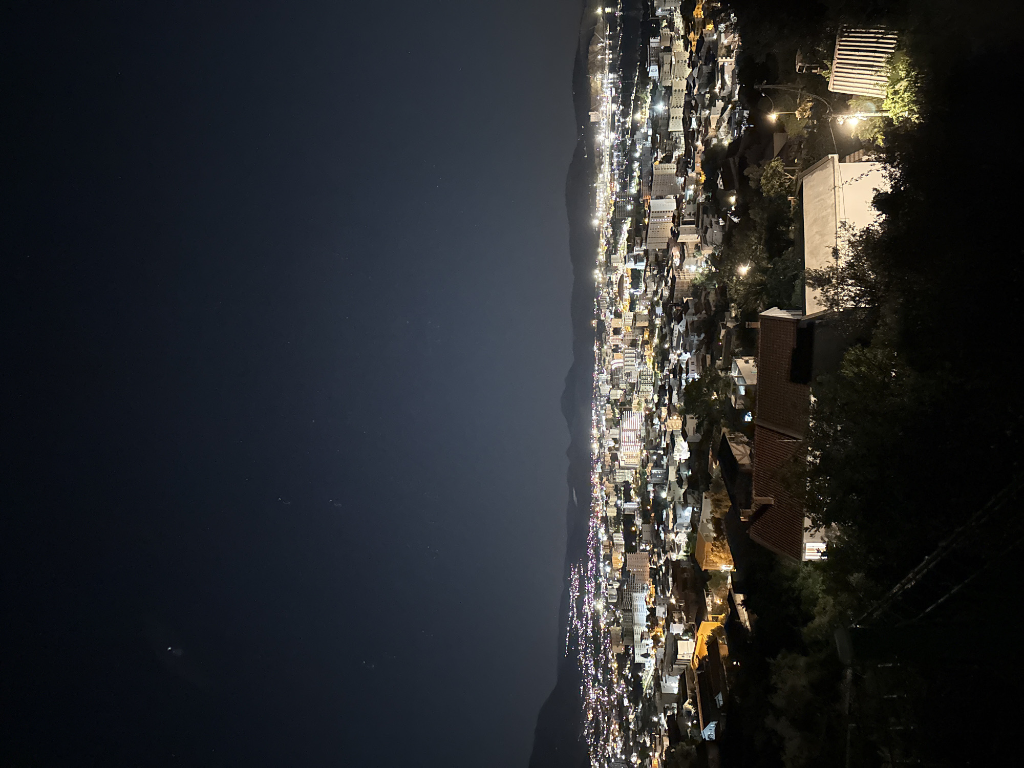 The image depicts a nighttime view of Šušanj, Montenegro. The right side of the image shows residential structures built on a hillside. Further to the left, a dark body of water is seen adjacent to the lights of the city. A mountain range sits between the water and the city lights. The time of day is night. The setting is urban.