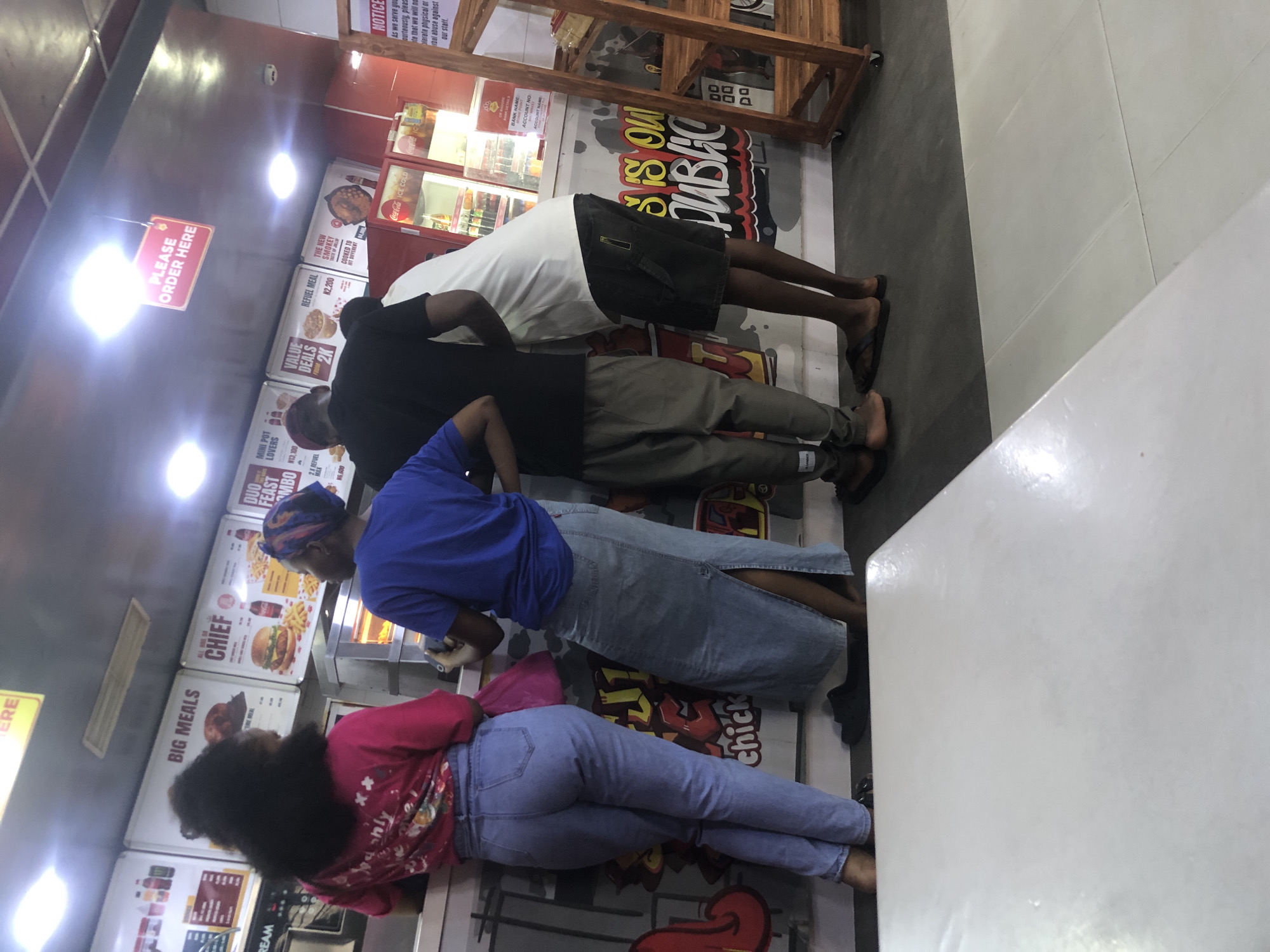 The image depicts the interior of a fast-food restaurant in Ado, Nigeria, likely a branch of "Chief" restaurant, as indicated by signage. Three individuals are visible from the mid-torso down, standing at a counter. The person on the left, wearing a pink top and blue jeans, is facing away from the camera. To their right, a person in a blue t-shirt and a long denim skirt is also facing away. The third person, to the far right, is wearing a black t-shirt and black shorts. Behind the counter, menus are displayed on the wall, featuring items like "BIG MEALS," "CHIEF," "DUO FEAST COMBO," and various deals with prices in Nigerian Naira. Refrigerated drink displays are also visible. The overall environment appears to be a casual dining establishment.