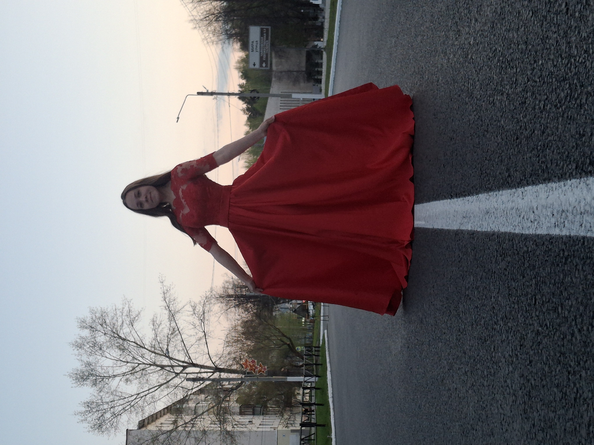 A woman in a red dress stands on a road in the city of Chaikovsky, Russia. The road has a white line painted on it. In the background, a sign reads "Mira Street" (Мира улица) in Russian. Also visible are trees, a building, a lamppost, and the sky, which appears to be dusk. The woman is holding up the sides of her dress with her hands.